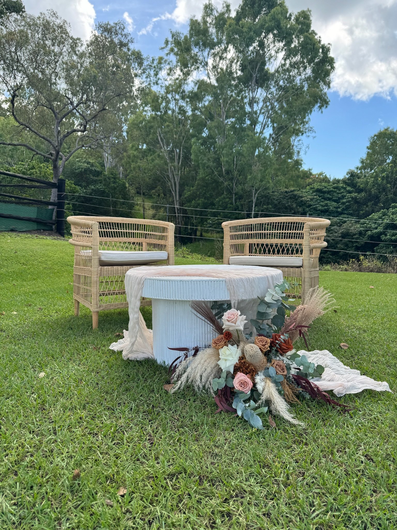 Outdoor setup with two wicker chairs and a round table draped with a white cloth, decorated with a floral arrangement of roses, eucalyptus, and dried plants on a grassy lawn, with trees and a partly cloudy sky in the background.
