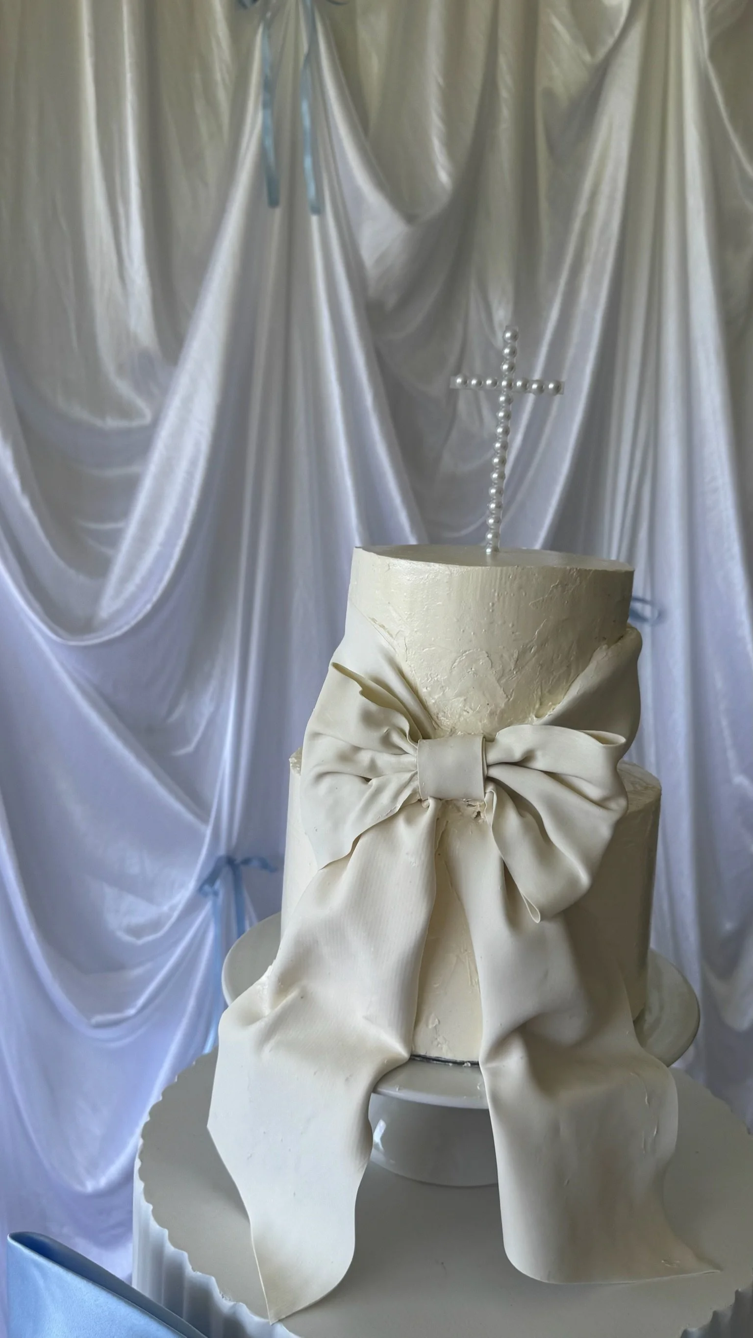 A white iced wedding cake with a large bow made of fondant or icing on the front, topped with a cross made of pearl-like beads, against a draped satin or silk fabric backdrop.