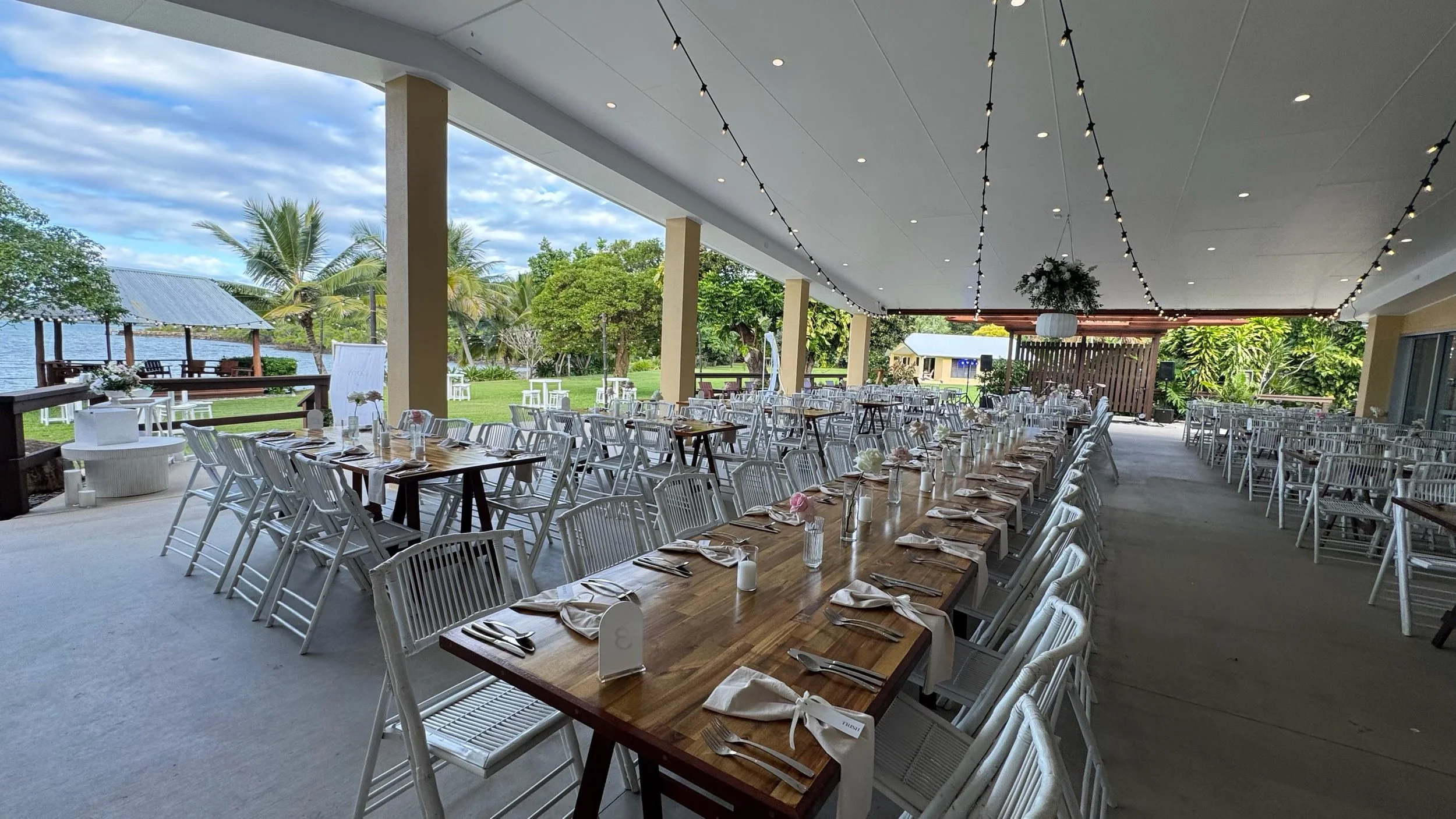 Indoor wedding or event space decorated with string lights, set for a meal with long wooden tables, white chairs, napkins, and tableware, with an open view of a grassy yard, palm trees, and a lake outside.