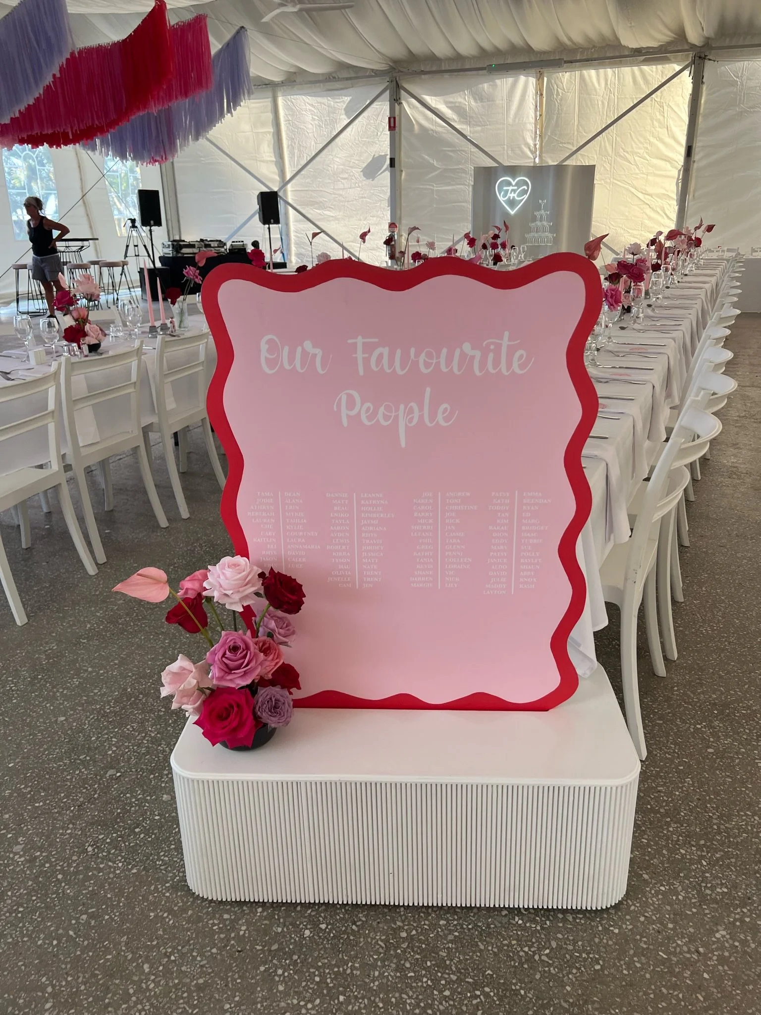 A decorated wedding reception area inside a tent with a pink and white theme. There are long tables with white tablecloths, pink and red flower centerpieces, and chairs. A large pink sign with the text 'Our Favourite People' and a list of names is pl