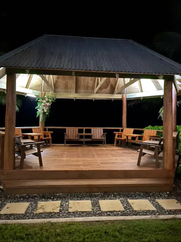 Wooden gazebo with benches and chairs, illuminated at night, with a pathway leading to it and decorative plants