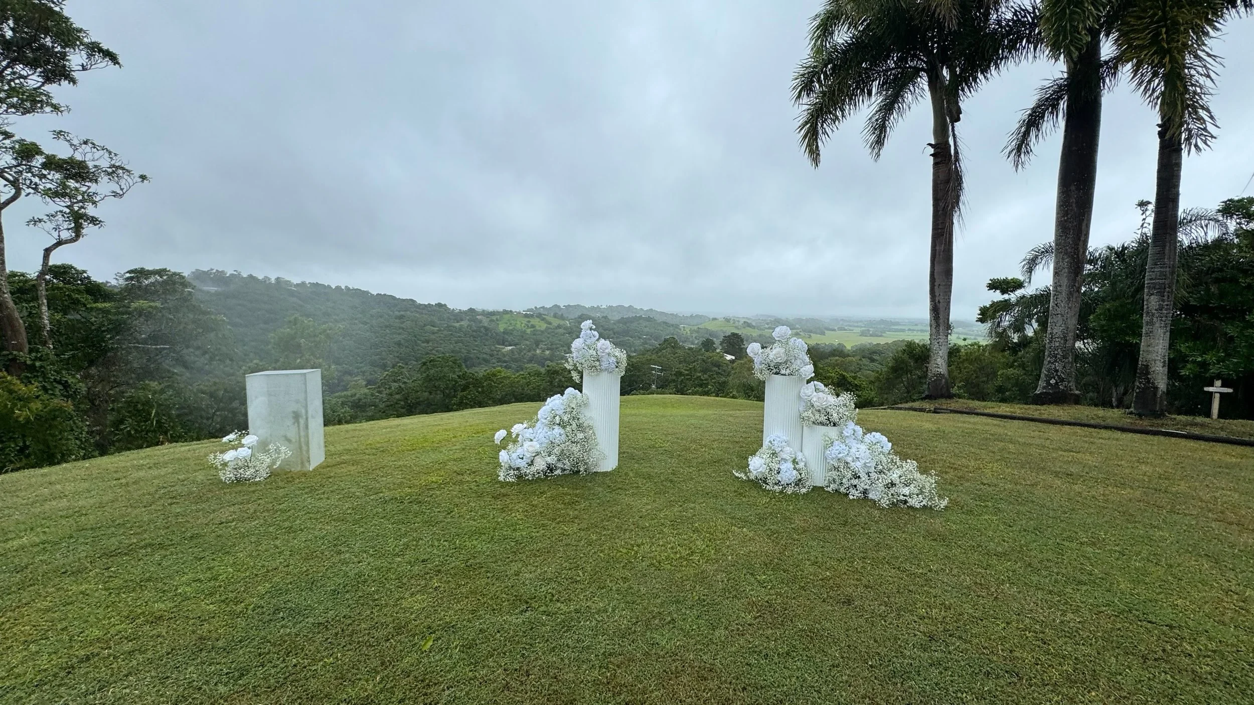 Outdoor wedding setup with white floral arrangements on tall white pedestals on a grassy hill, with trees and a cloudy sky in the background.