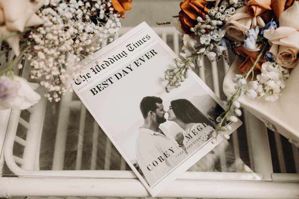 A newspaper titled "The Morning Times" with a headline "Best Day Ever" is surrounded by flower arrangements on a white chair.