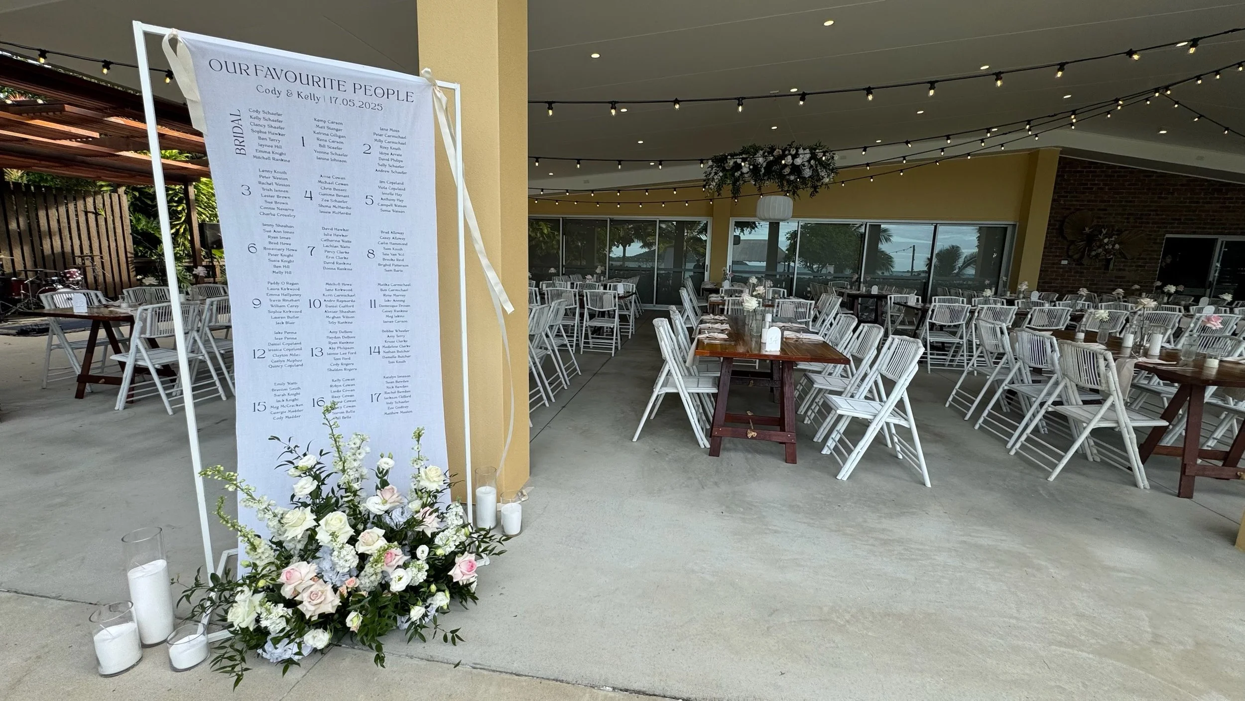 Wedding reception setup with white chairs and tables, floral arrangements, and a seating chart board on the left side with the names of guests for Cody and Kelly's wedding on May 17, 2025.