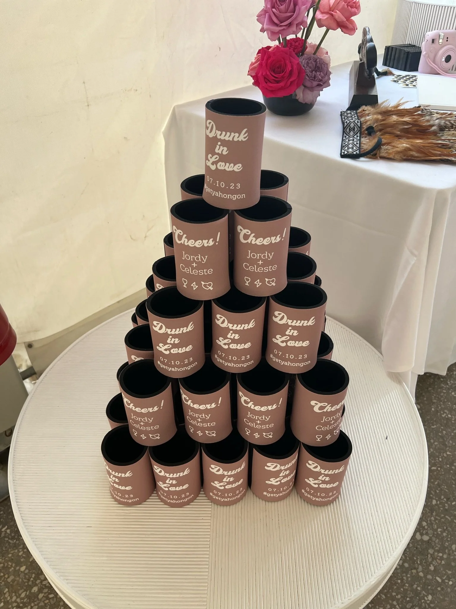 A pyramid of pink drink cans with personalized labels on a round table, with a table in the background holding a bouquet of pink and purple roses, a camera, and a feathered accessory.