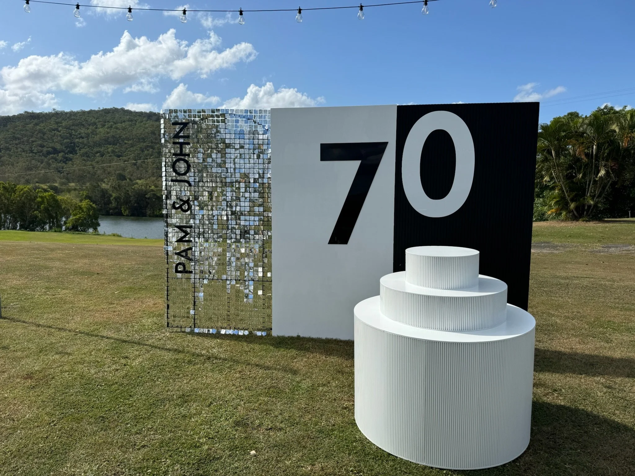 Outdoor display with the number 70, a reflective mirrored panel with 'PANTONE & JON' written on it, and a white tiered cylindrical structure on grass with trees and a lake in the background.