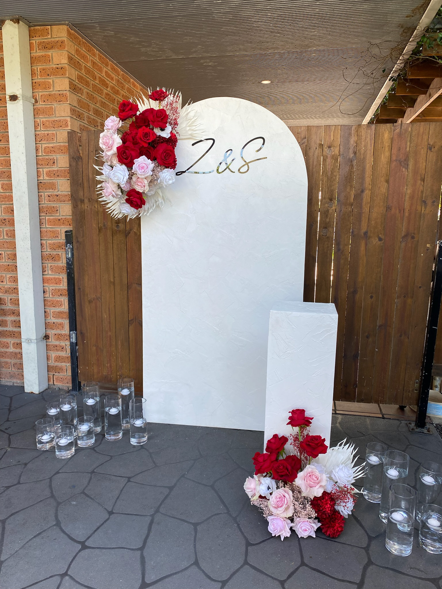 Wedding or event backdrop with a white textured curved board featuring the initials 'Z & S' in black and gold, decorated with red, pink, and white roses, with candles in glass holders on the ground.