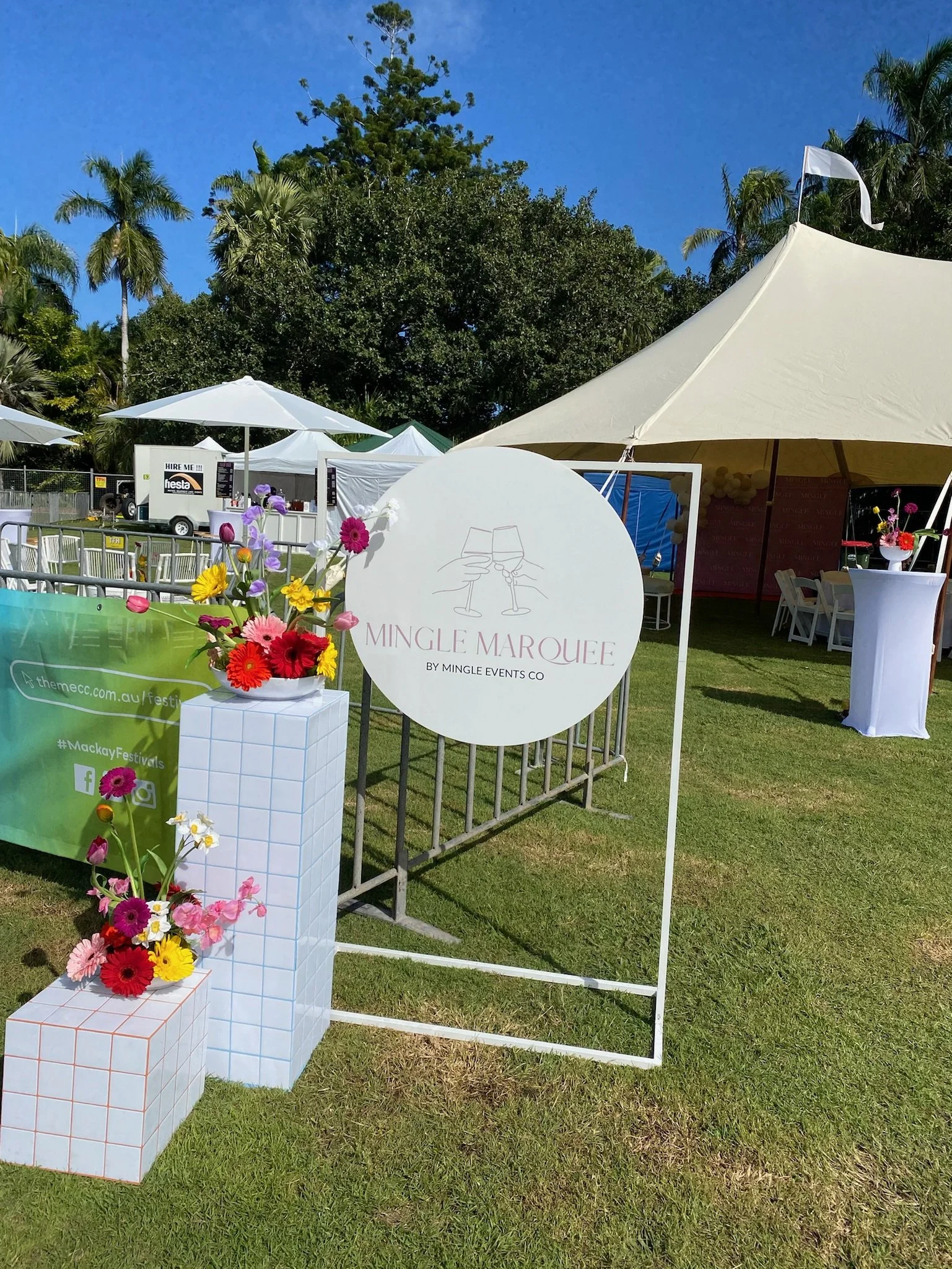 Outdoor event setup with white tents, umbrellas, and a sign that reads 'Mingle Marquee by Mingle Events Co,' decorated with colorful flowers.