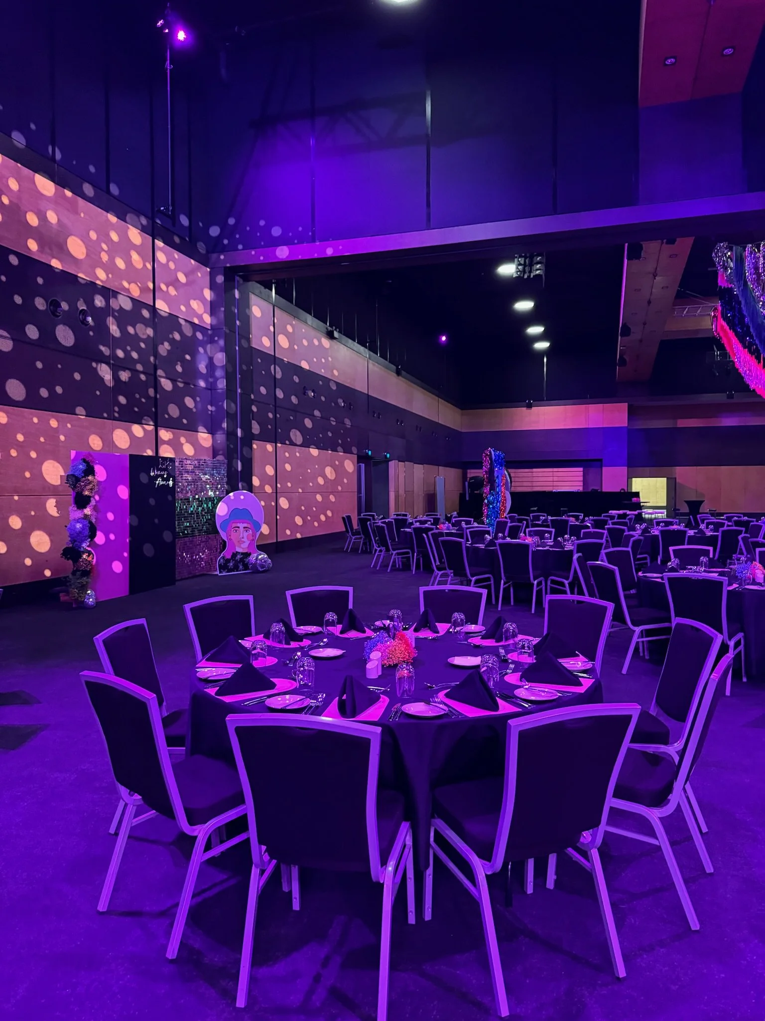 Event hall decorated with purple lighting, round tables with purple tablecloths, black napkins, glassware, and floral centerpieces, prepared for a formal event or celebration.