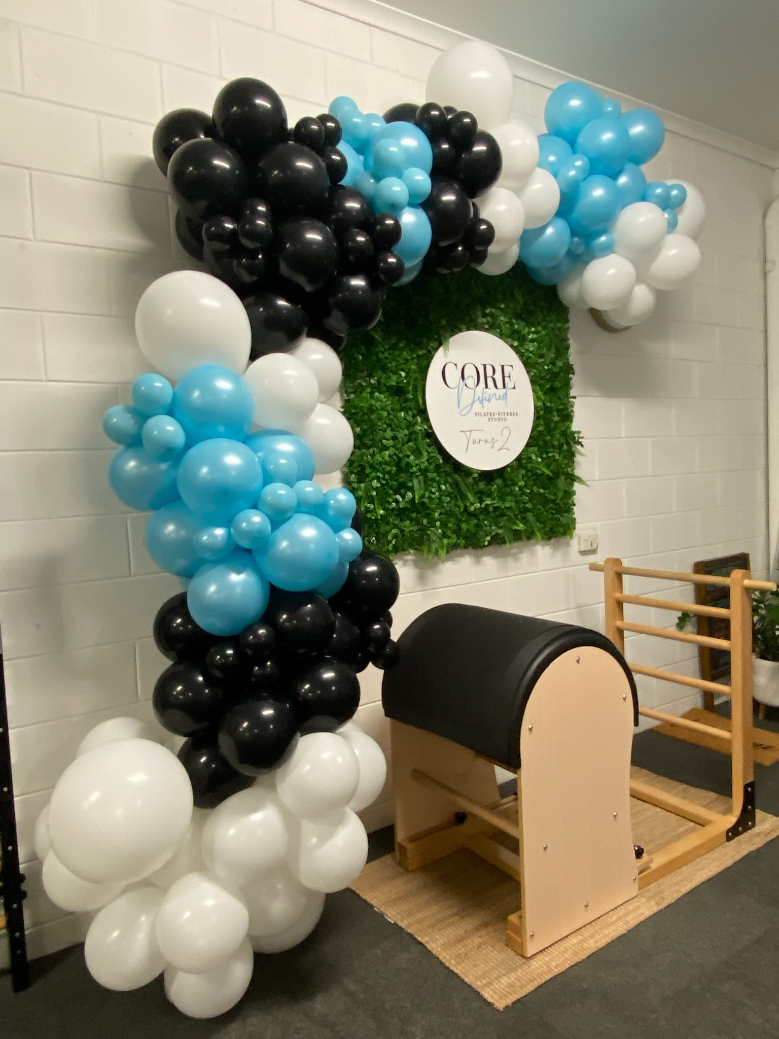 Balloon arch with black, white, and light blue balloons surrounding a green foliage wall with a round sign reading 'CORE Pilates + Fitness Studio Turn 2' inside a fitness studio with a Pilates reformer.