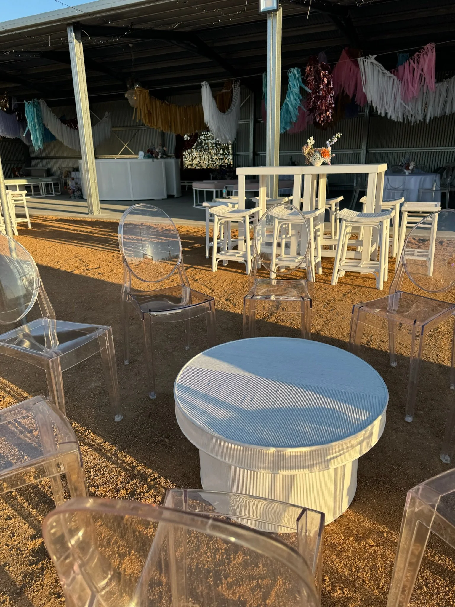 Decorated outdoor event space with white furniture, clear chairs, and pastel-colored paper garlands hanging from the ceiling, illuminated by warm sunlight.