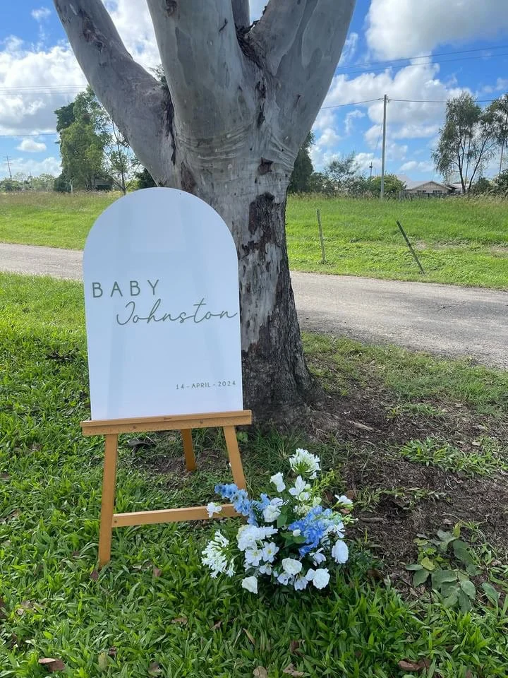 A white arched sign on a small wooden easel memorializes a baby named Johnston born on April 14, 2024, placed next to a tree with a bouquet of white and blue flowers at its base, outdoors on green grass with a gravel road and trees in the background.