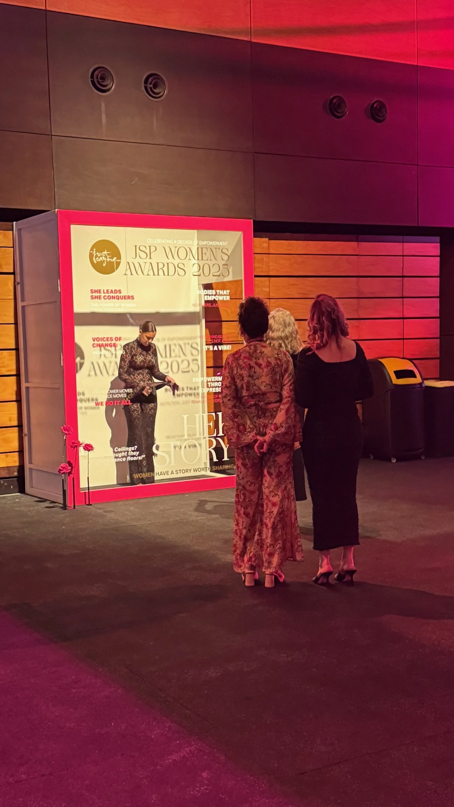 Three women standing in front of a large sign at the JSP Women's Awards 2023 event, with colorful lighting in the background.