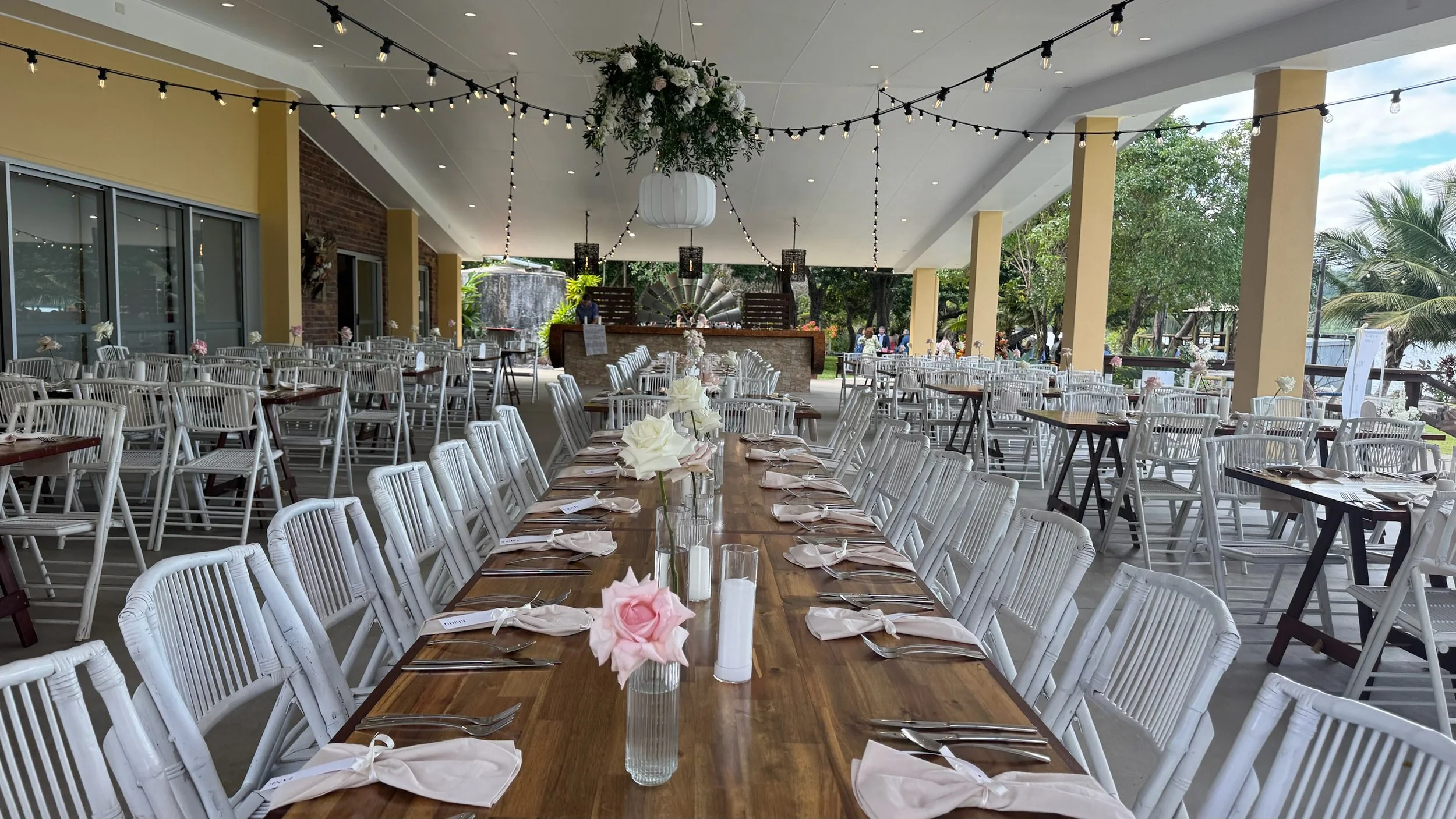 Decorated outdoor event space with long wooden tables, white chairs, flower centerpieces, and string lights, set up for a celebration.