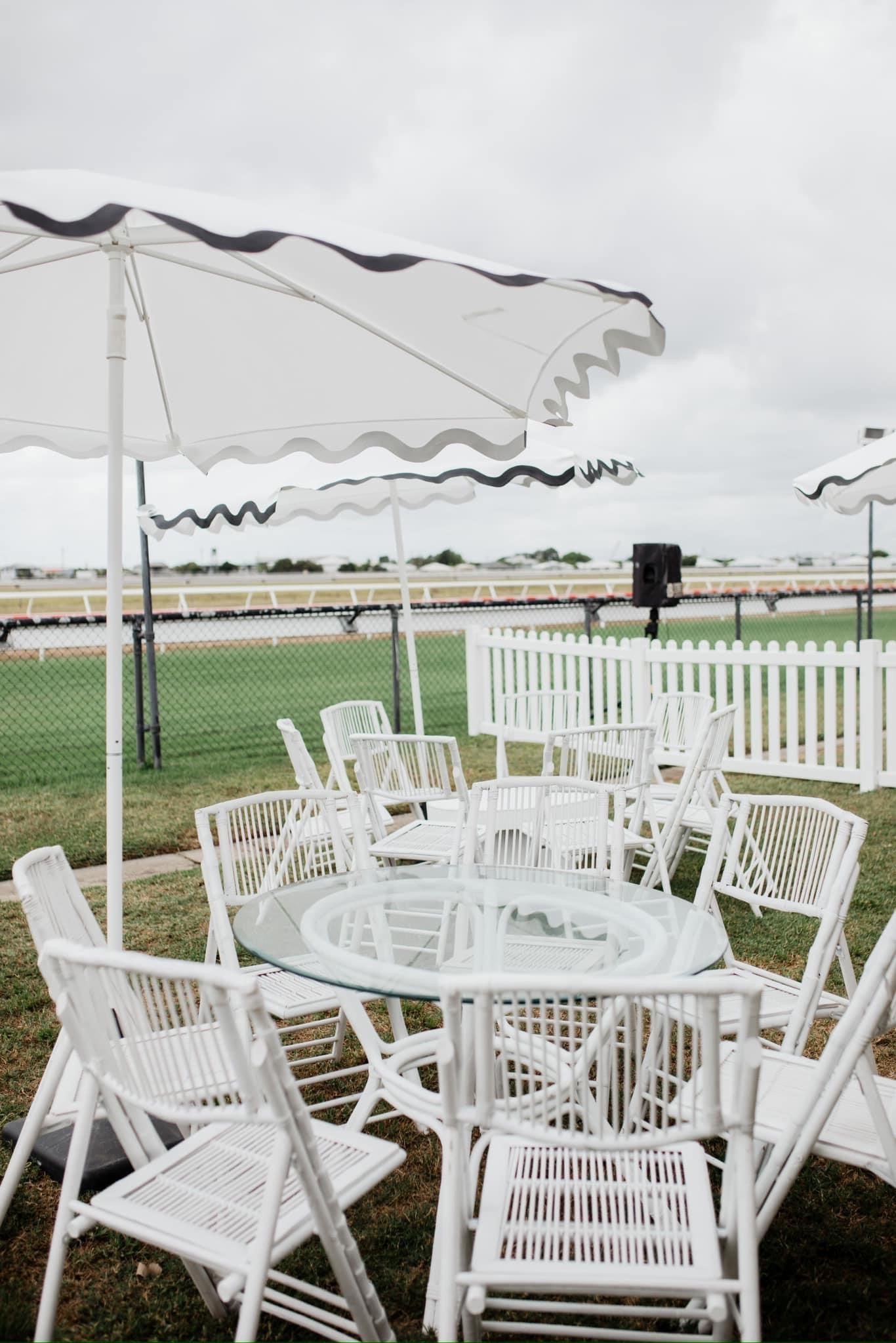 Empty white outdoor table with glass top surrounded by white chairs, with large white umbrellas overhead, at a grassy area near a racetrack.