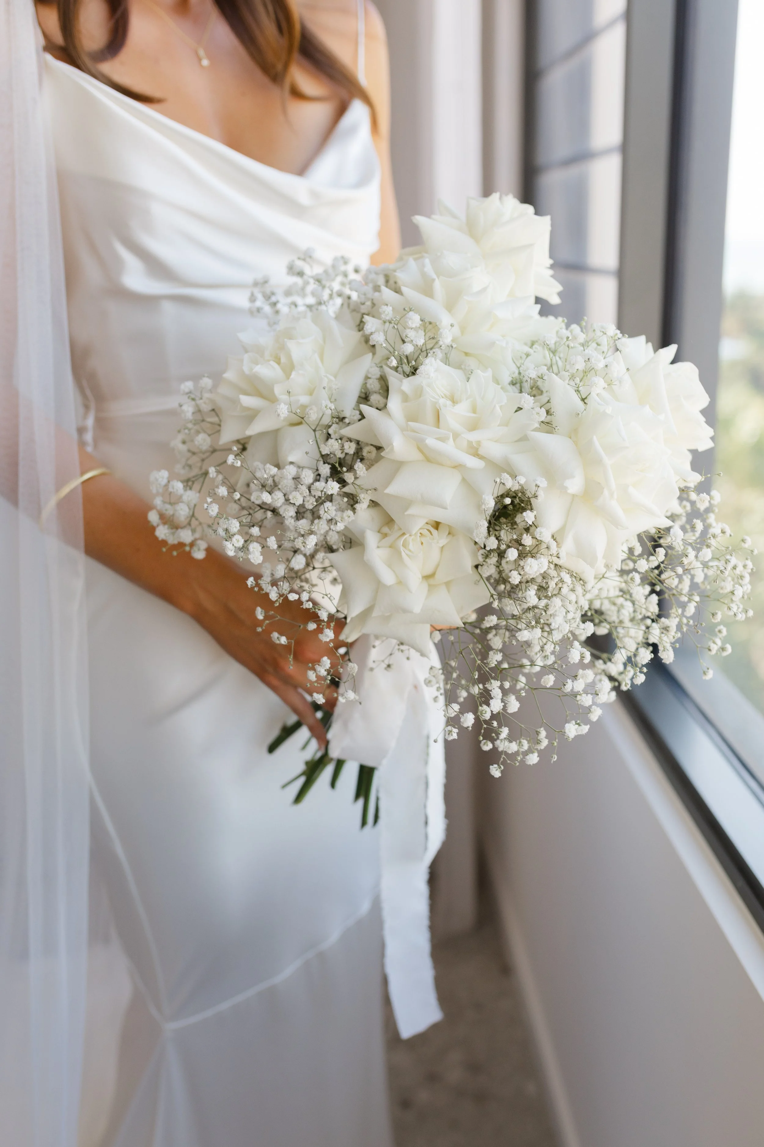 Bride holding a bouquet of white roses and baby's breath, wearing a white satin wedding dress and a gold bracelet, standing by a window.