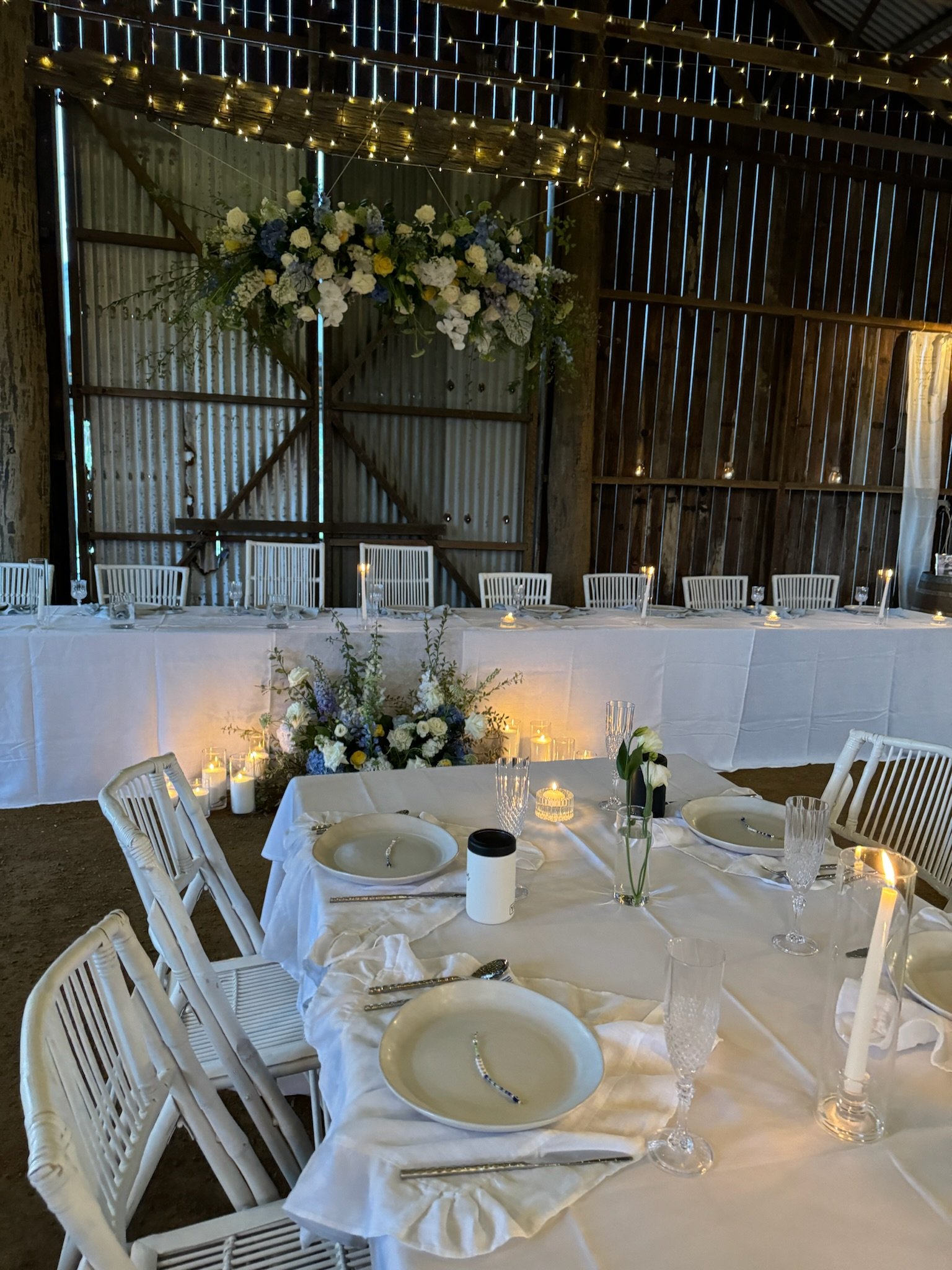Indoor rustic wedding reception with a long head table decorated with white flowers and candles, a round table set with plates, silverware, and glasses, and a backdrop of string lights and a floral arrangement.
