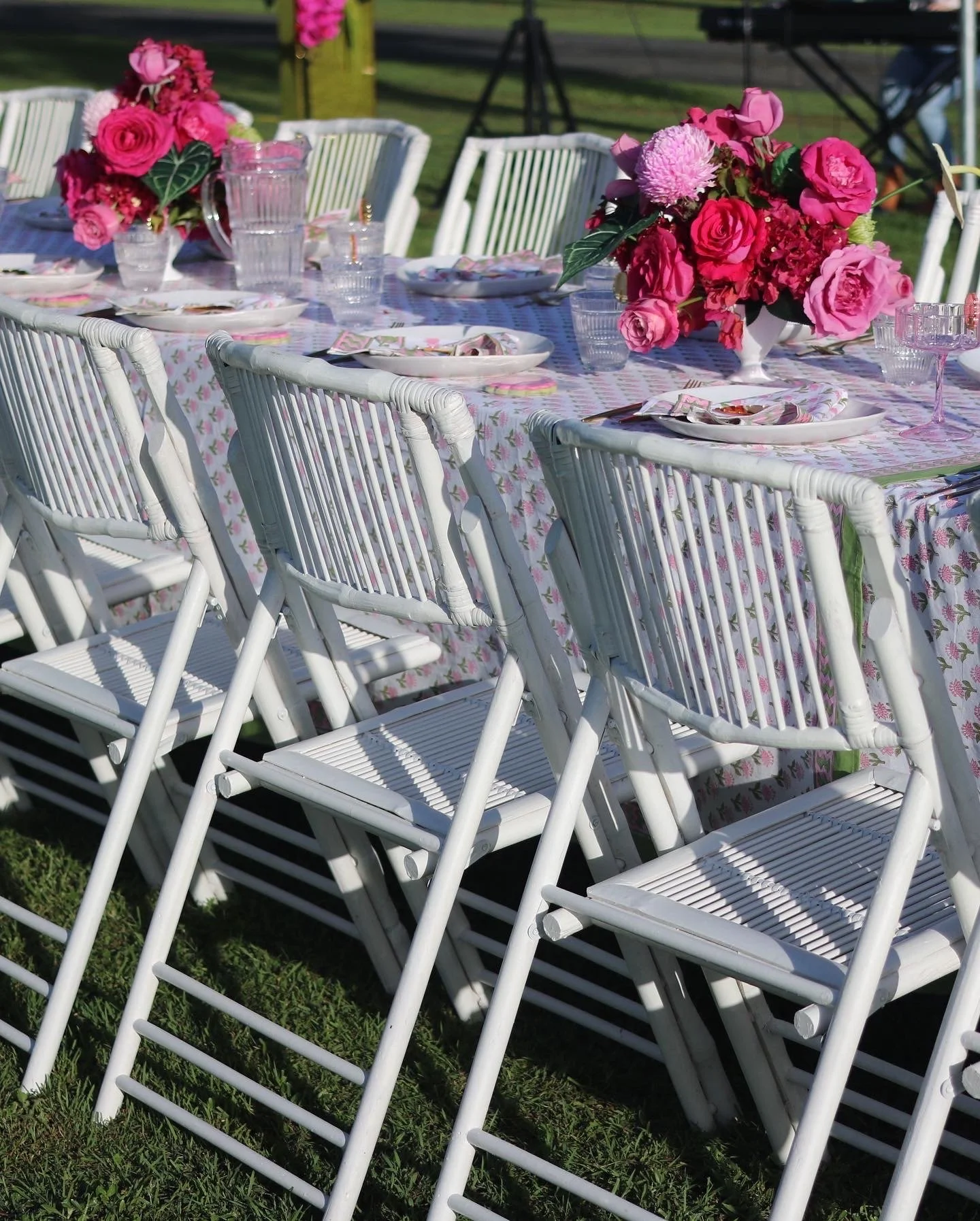 An outdoor table set with pink and red floral centerpieces, white plates, glasses, and napkins, surrounded by white chairs with woven seats and backs, on a grassy area with a tent in the background.