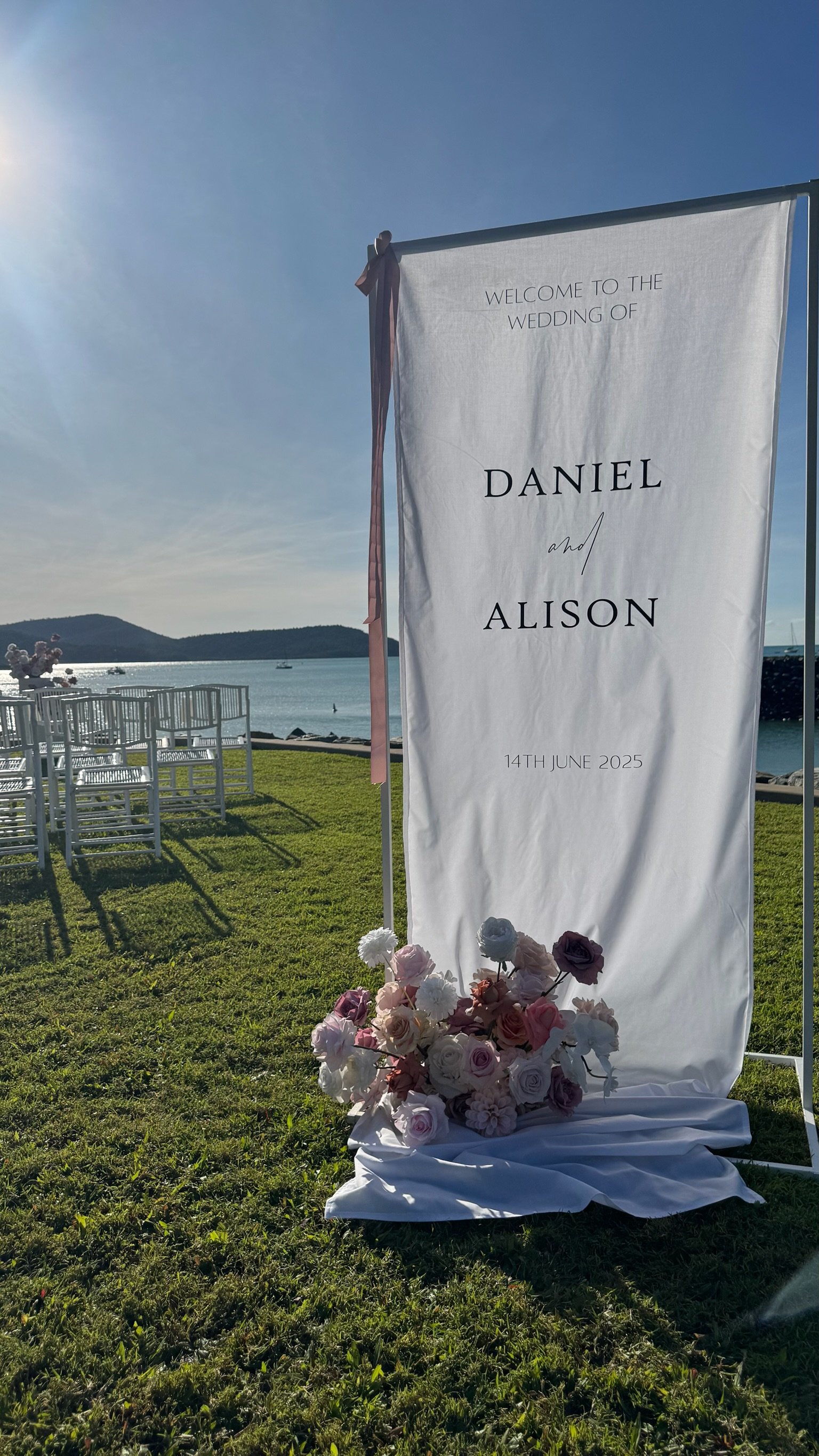 Wedding welcome banner for Daniel and Alison by the water on June 14, 2025, decorated with pink and white flowers, and ribbons.