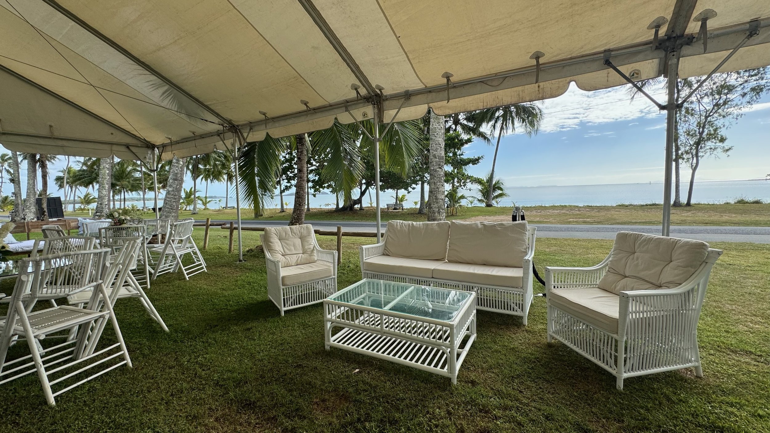 Outdoor seating area with white wicker furniture, cushioned chairs, a glass-topped table, and a large canopy, set near a tropical beach with palm trees and ocean in the background.