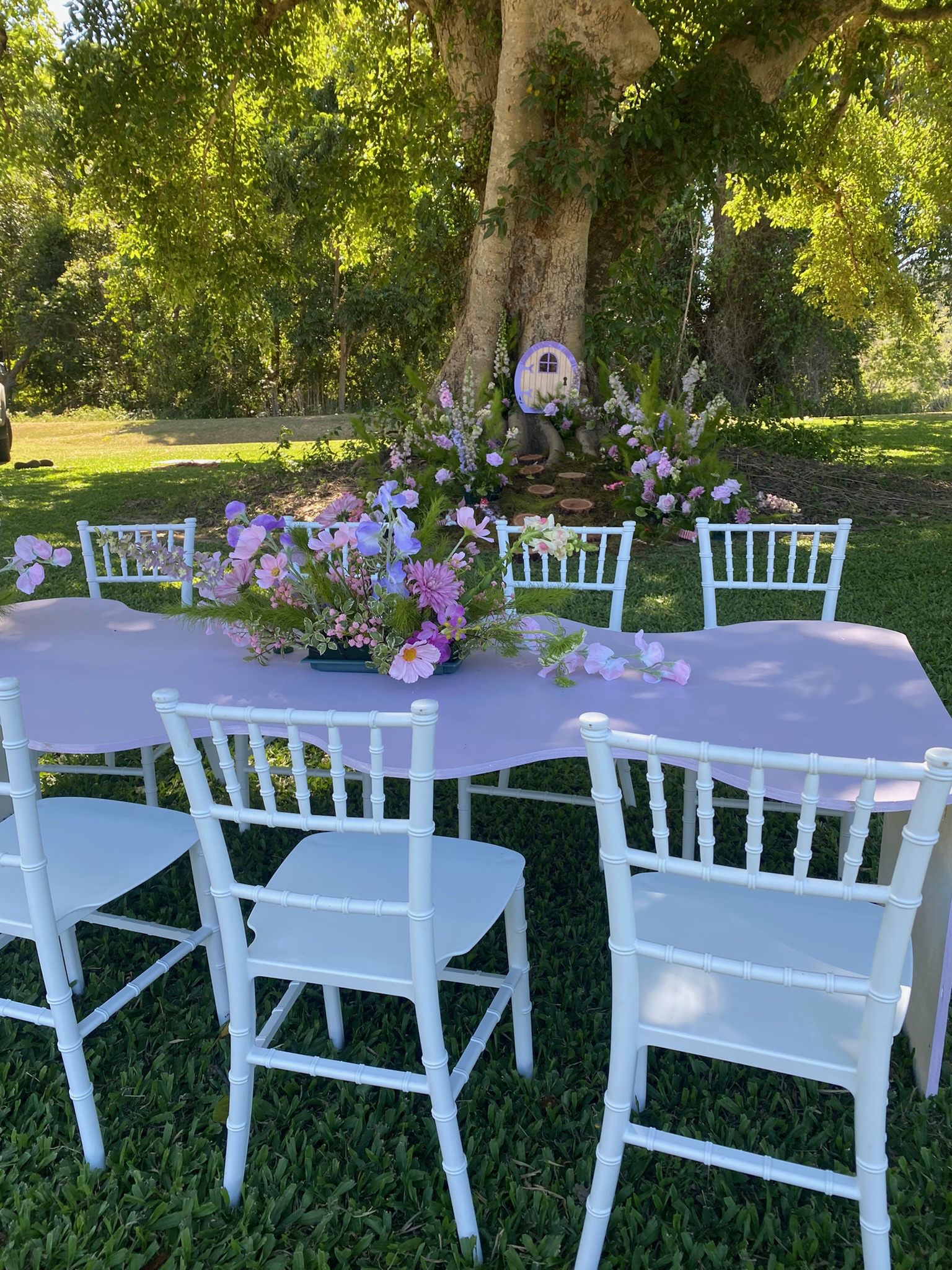 Outdoor table decorated with a pink tablecloth and a floral centerpiece, surrounded by white chairs, set in a garden with a large tree and floral arrangements in the background.