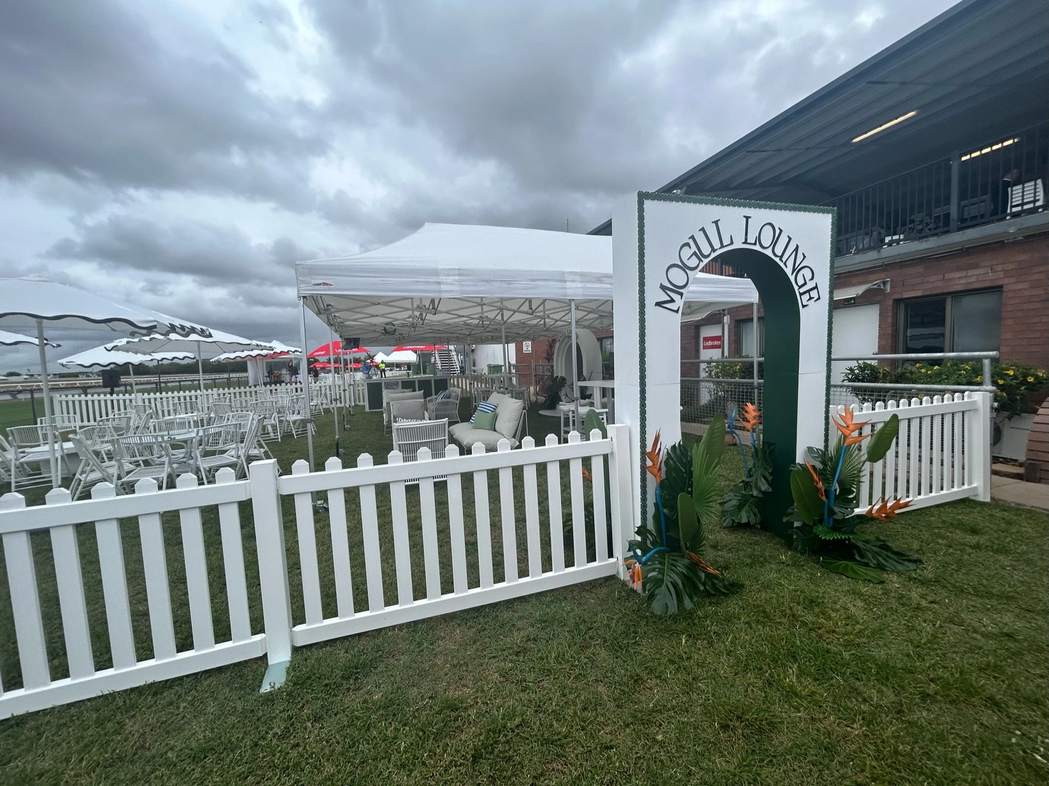 Outdoor seating area with white chairs, tables, and tents, surrounded by a white picket fence, under cloudy skies, at Mogull Lounge with decorative tropical plants at the entrance.