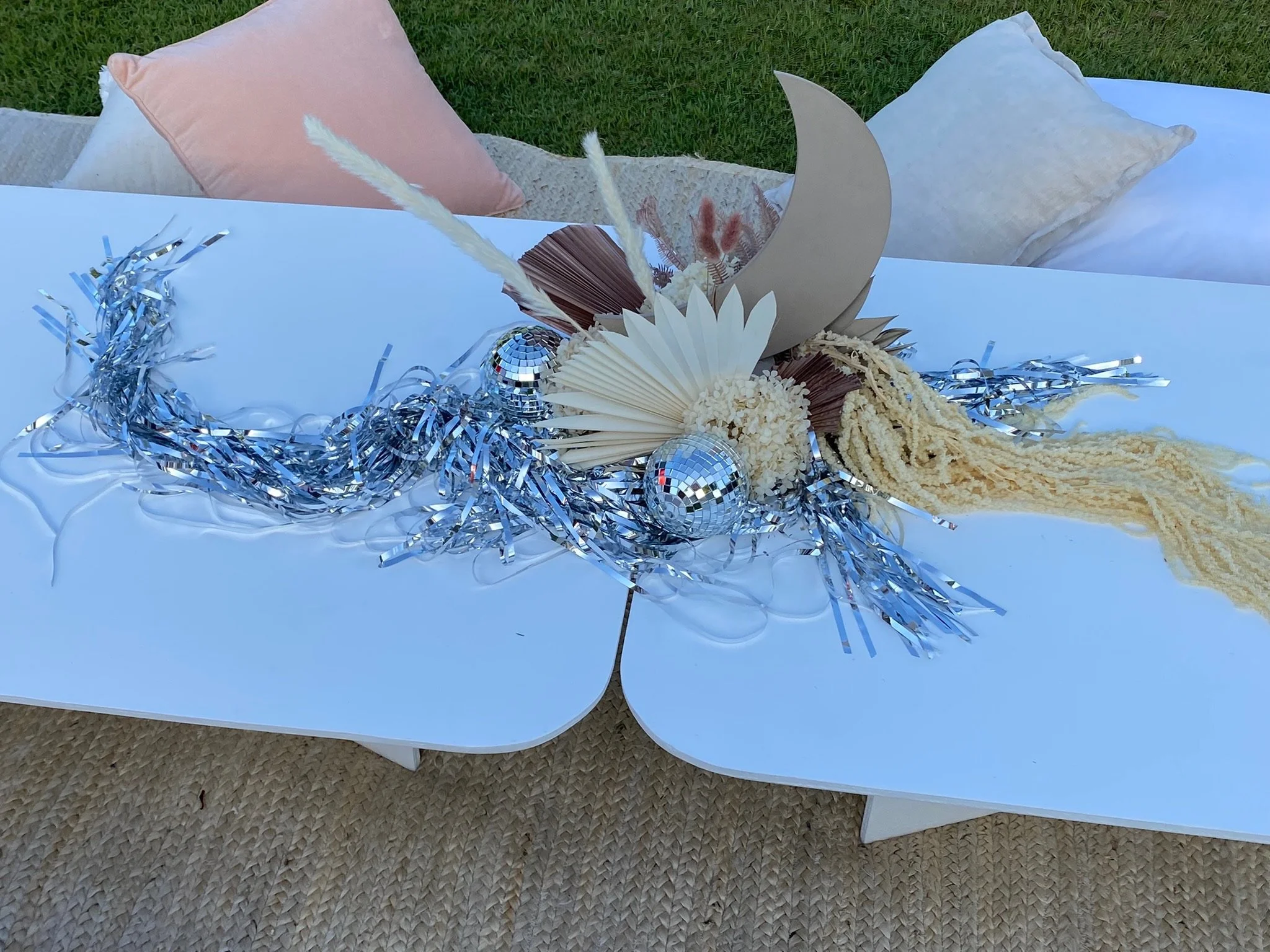 Decorative centerpiece with silver tinsel, disco balls, paper fans, and dried flowers on a white table, outdoor setting with cushions in the background.