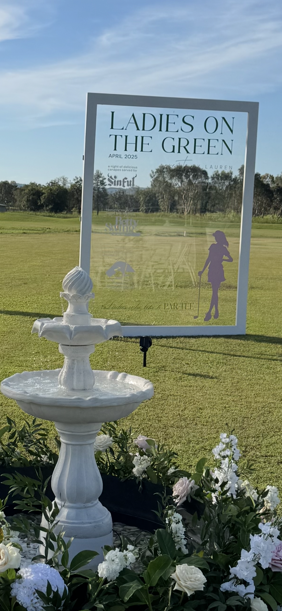 A decorative white fountain on a grassy golf course in front of a sign that reads 'Ladies on the Green April 2025' with an illustration of a woman, along with some white and pink flowers at the base of the fountain.