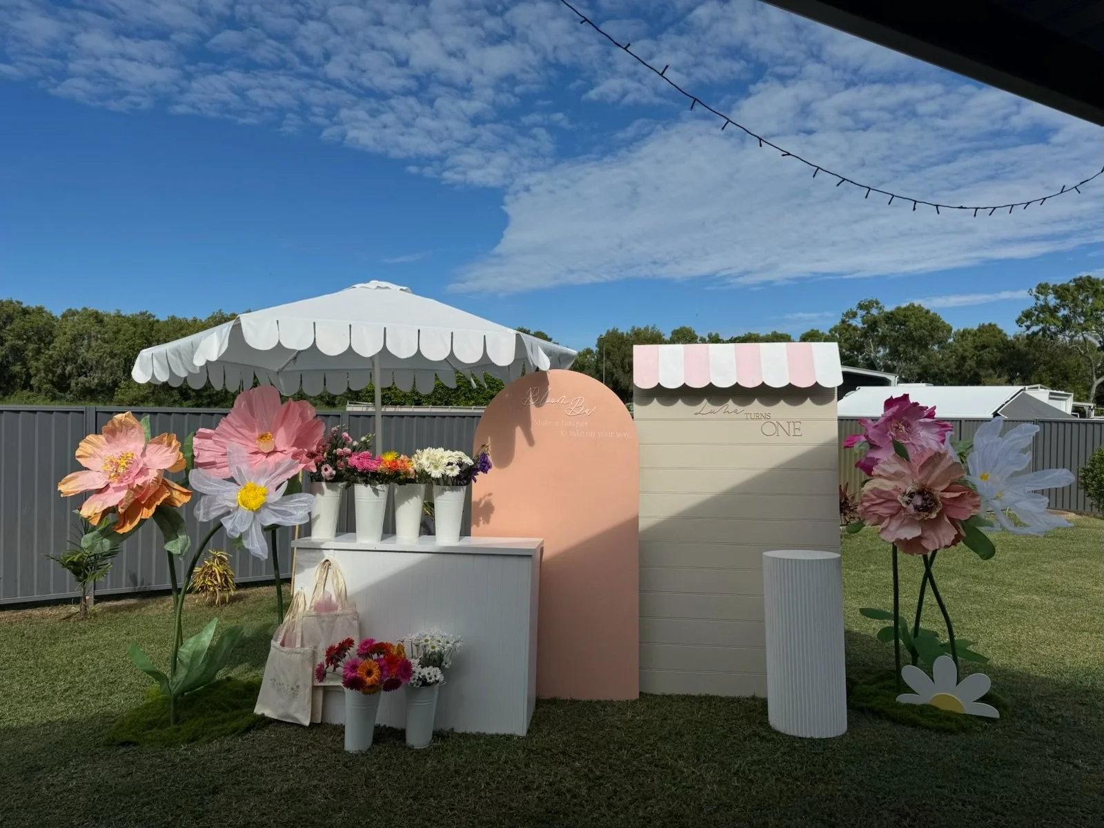 Outdoor pink and white birthday party setup with large flower decorations, a pink backdrop, and a white umbrella with string lights overhead on a grassy yard.