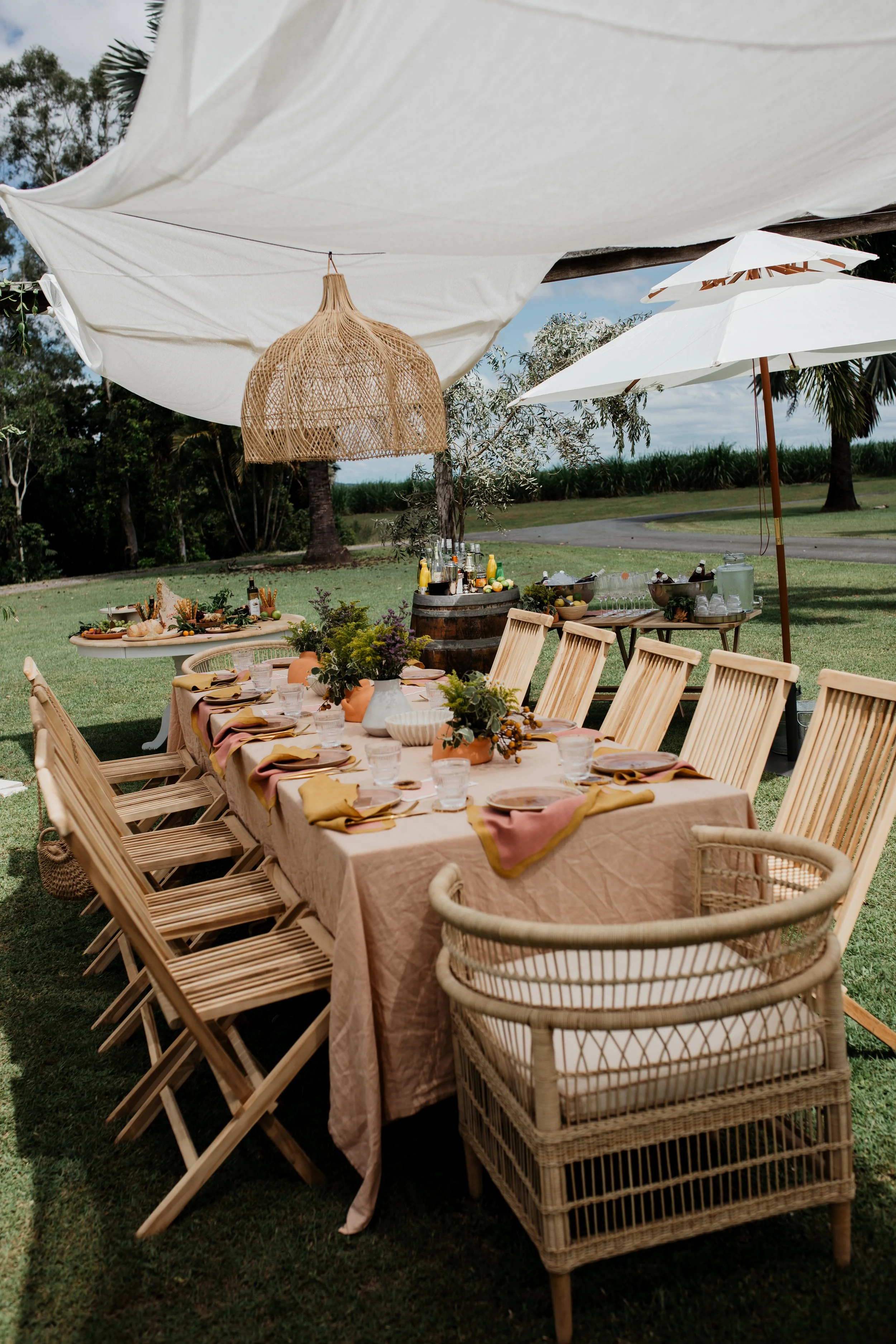 Outdoor dining table set up with beige tablecloth, flower arrangements, and place settings on a lush lawn under large white shade umbrellas and parasols, with trees and a clear sky in the background.