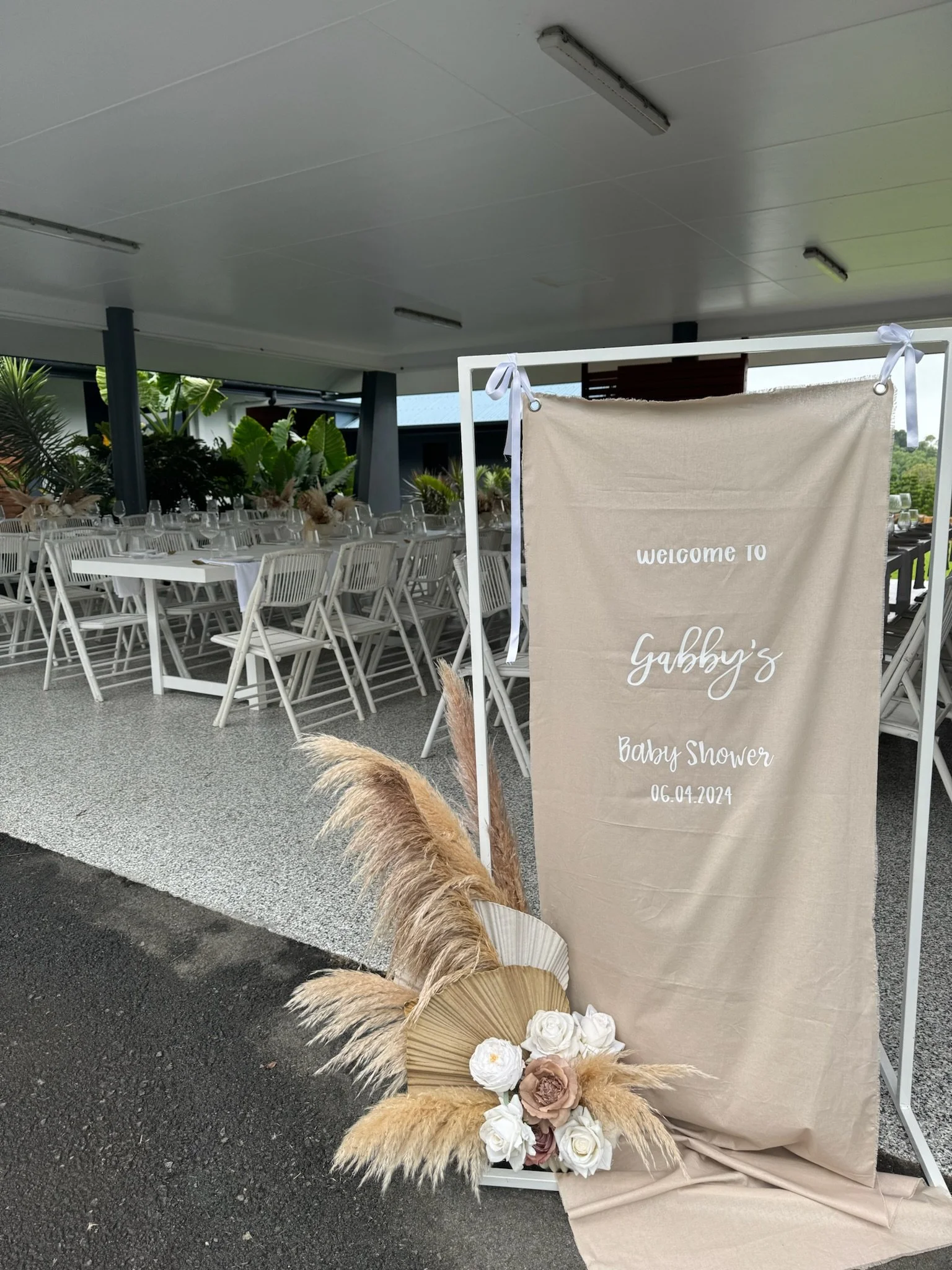 Baby shower celebration setup with a welcoming banner for Gabby, decorated with flowers and pampas grass, at an outdoor venue with tables and chairs.