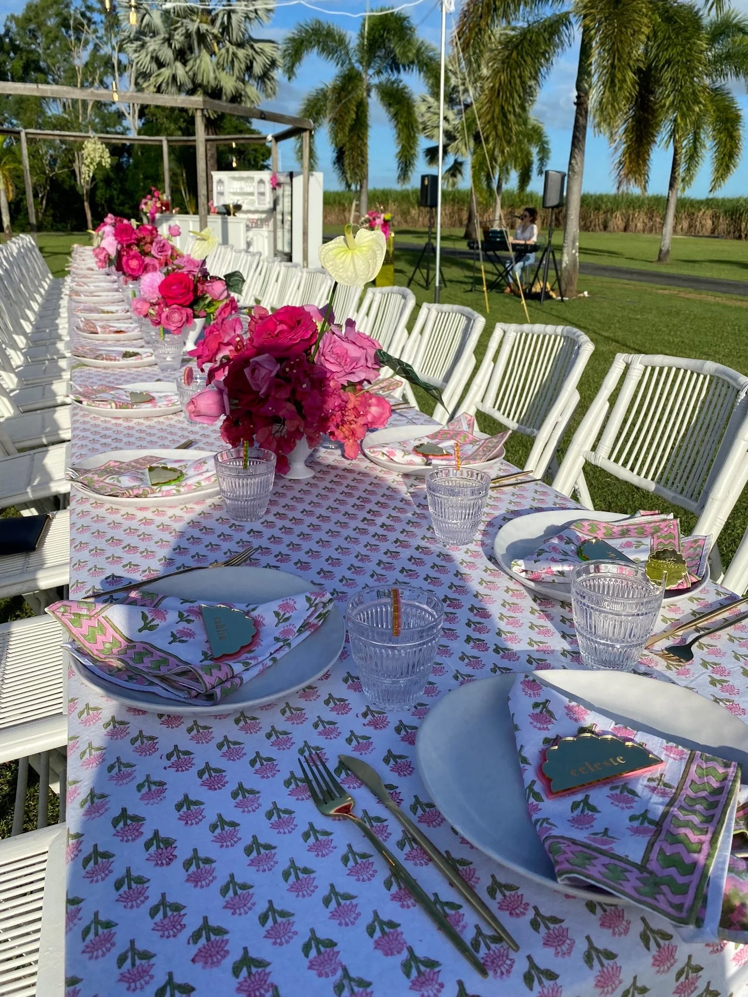 Outdoor dinner table set with pink floral centerpieces, pink and white napkins, gold cutlery, and clear glasses, surrounded by white chairs, in a lush green garden with palm trees, a blue sky, and a musician playing a keyboard in the background.