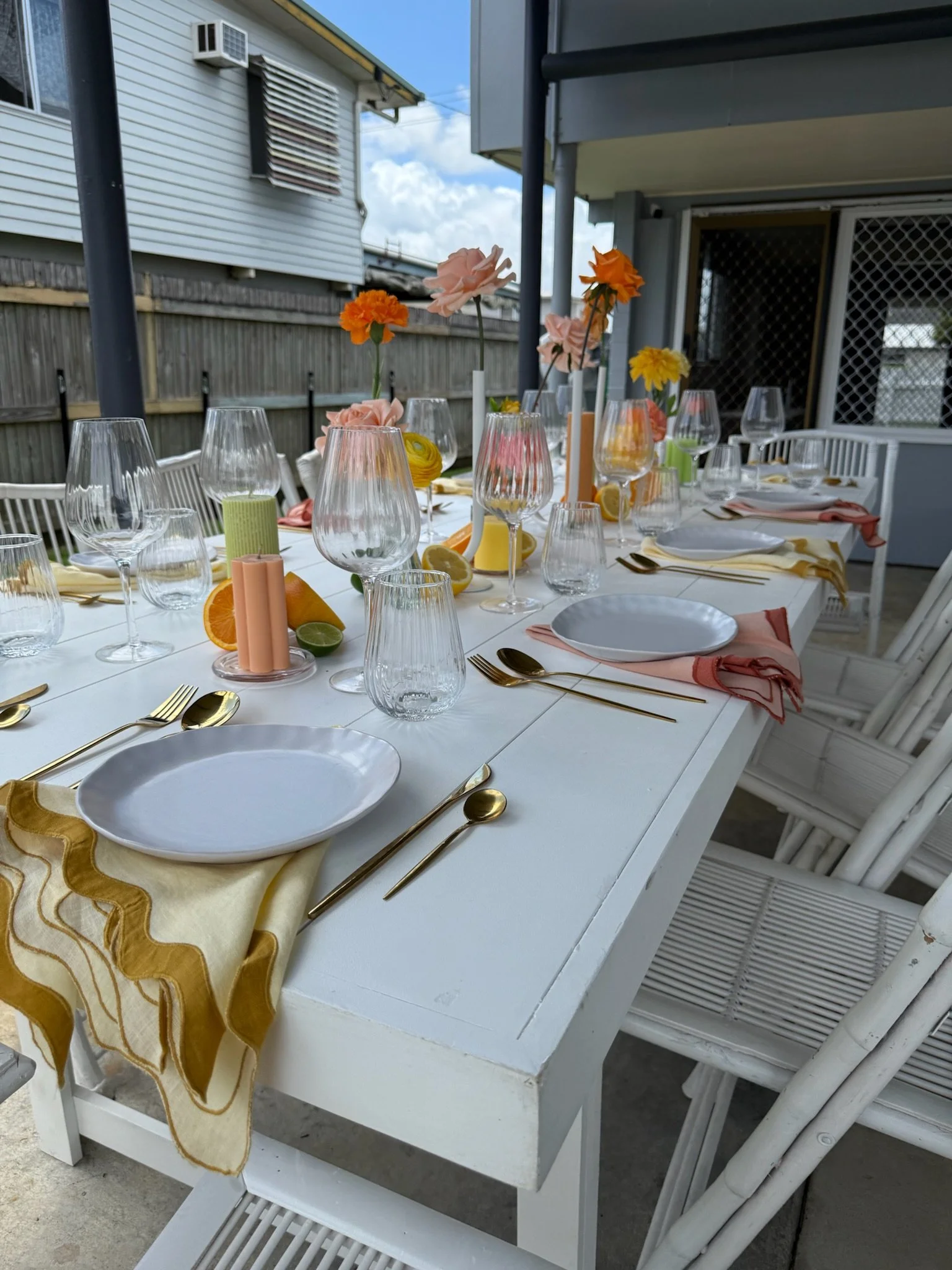 A long outdoor dining table decorated with pastel-colored flowers in vases, citrus fruit slices, candles, and set with white plates, gold flatware, and cloth napkins on a white tablecloth, under a covered patio.