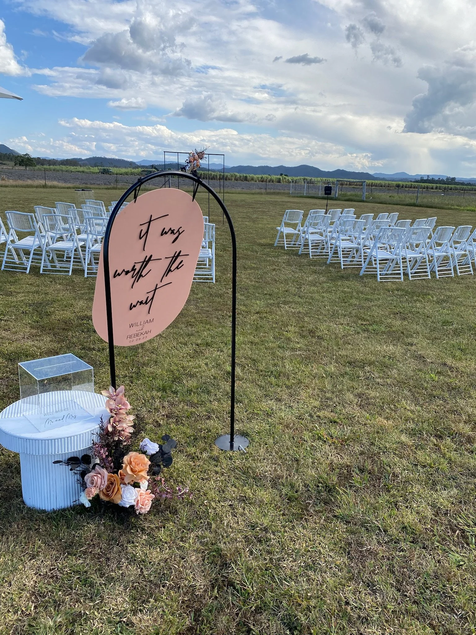 Outdoor wedding setup with white folding chairs arranged in rows on a grassy field, a decorative sign reading 'Twas WORTH the wait' on a pink oval board, flower arrangements at the base of the sign, and a flower arch in the distance, with mountains a