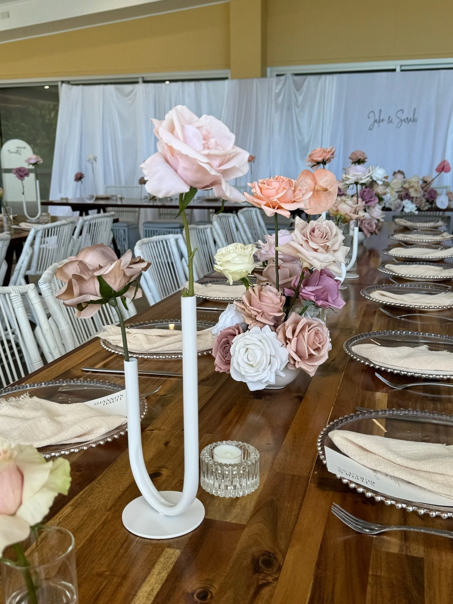 Elegant dining table decor with pink and white roses in a vase and a white modern candle holder with a candle, set on a wooden table with silver chargers and beige napkins, prepared for a formal event.
