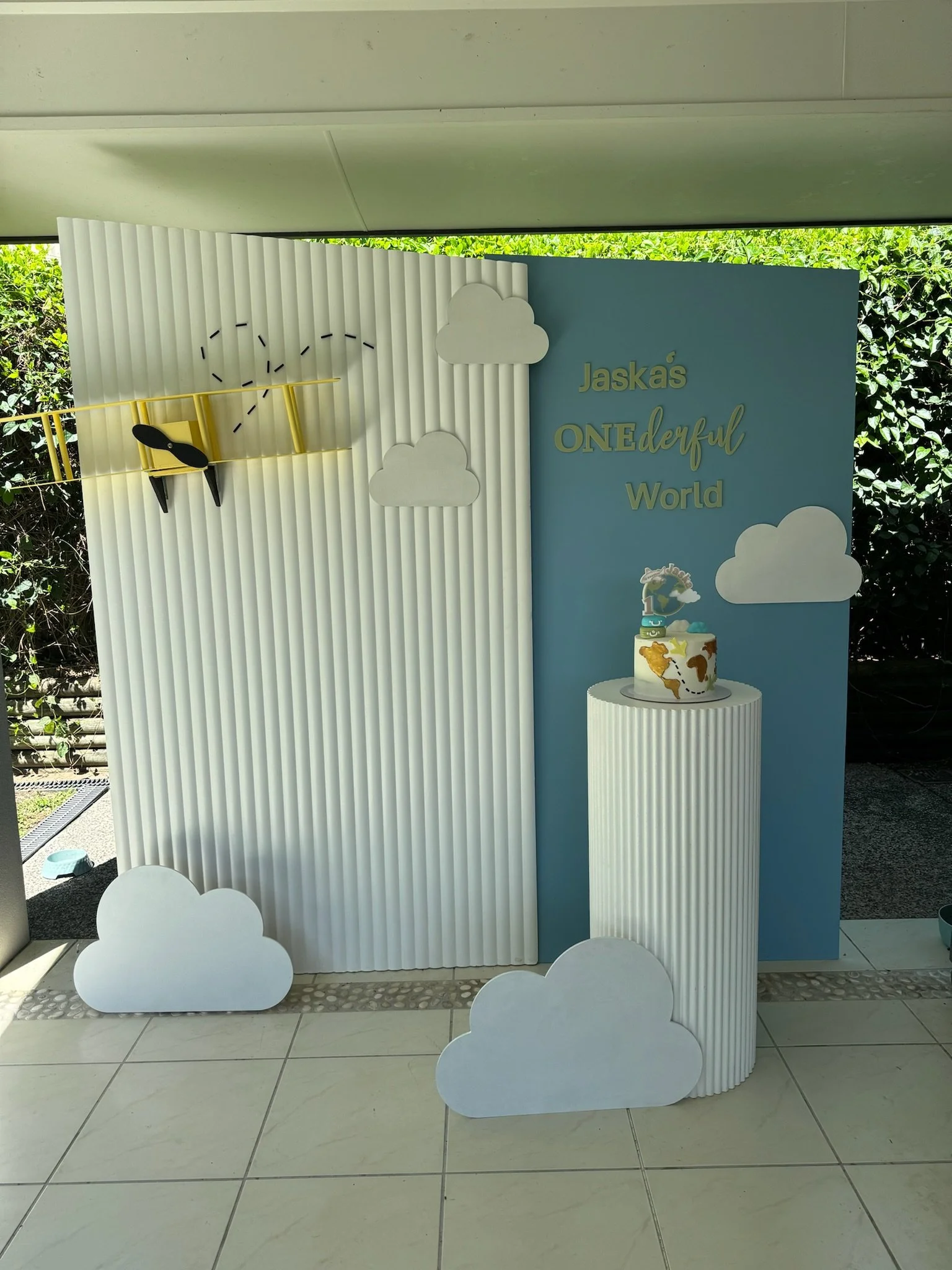 Decorative backdrop for a child's birthday party featuring clouds, a fishing pole with a fish, and a cake with a unicorn topper and colorful giraffe pattern, with a sign reading 'Jaskas ONEderful World'.