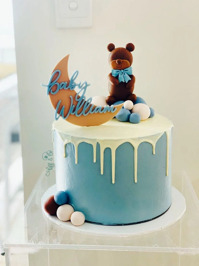 A cake with blue and white frosting, topped with a teddy bear figure, a moon-shaped topper with the words 'Baby William', and small blue, white, and brown spherical decorations.