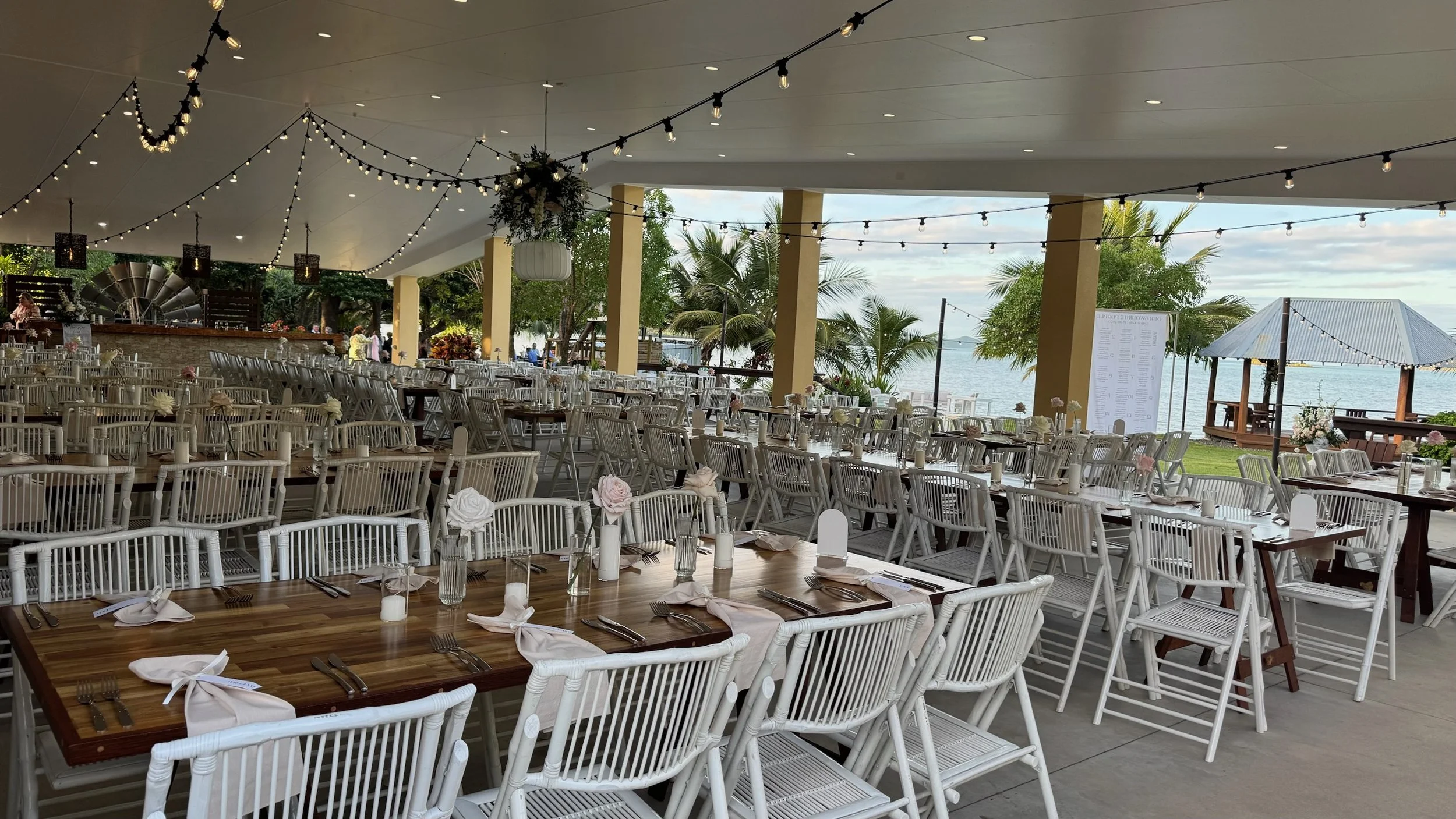 A spacious outdoor event space decorated with string lights and white chairs, overlooking a tropical landscape with palm trees and water in the background.