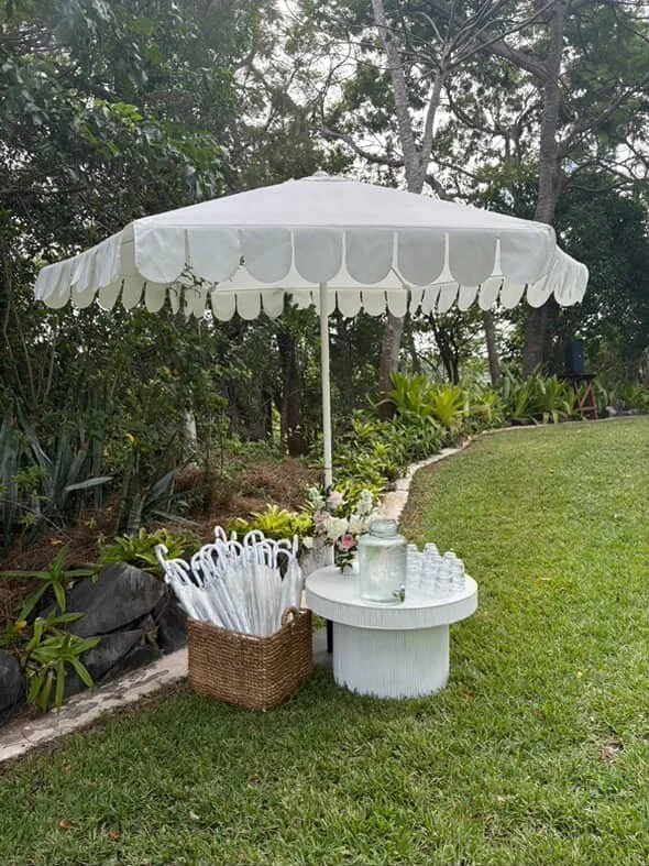 A white outdoor table with a matching umbrella, set up in a grassy garden area. The table holds a flower arrangement, a large glass jar, and several small glasses. Nearby, a wicker basket contains clear umbrellas. The background features lush greener