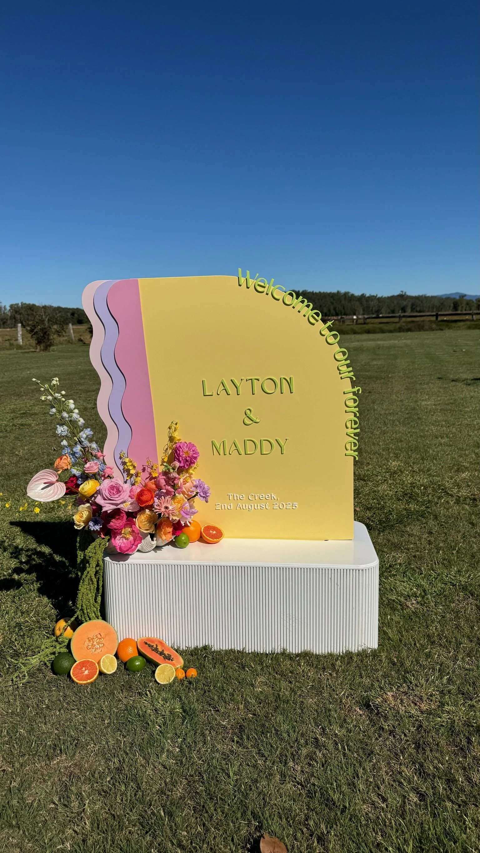 Decorative outdoor wedding sign with colorful flowers, artificial fruits, and pastel-colored wavy design, welcoming guests for Layton and Maddy's wedding on August 2, 2025.