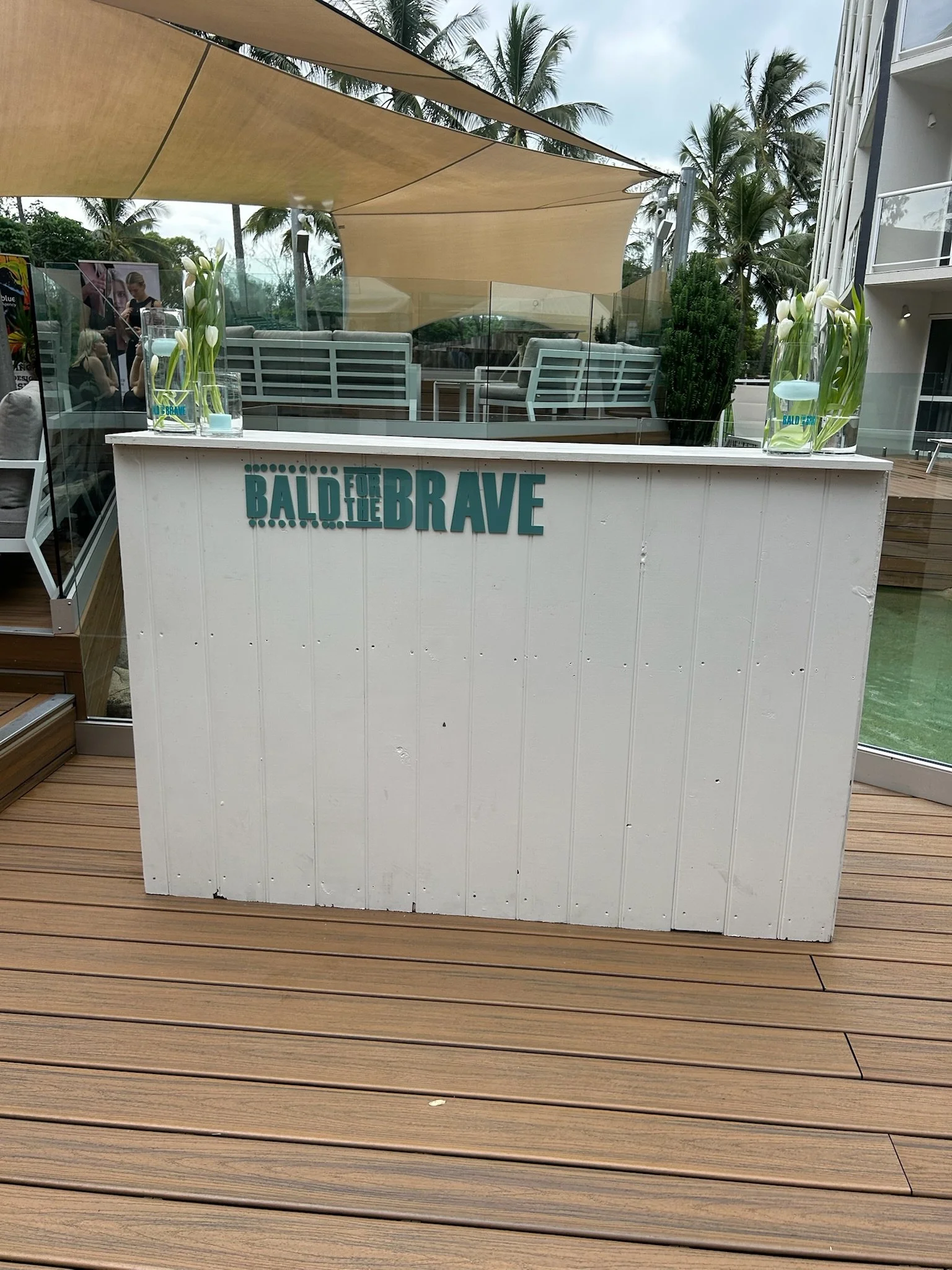 Outdoor bar with a white front, decorated with teal lettering that reads 'BALD FOR THE BRAVE', with two vases of white flowers on top and surrounded by tropical palm trees.