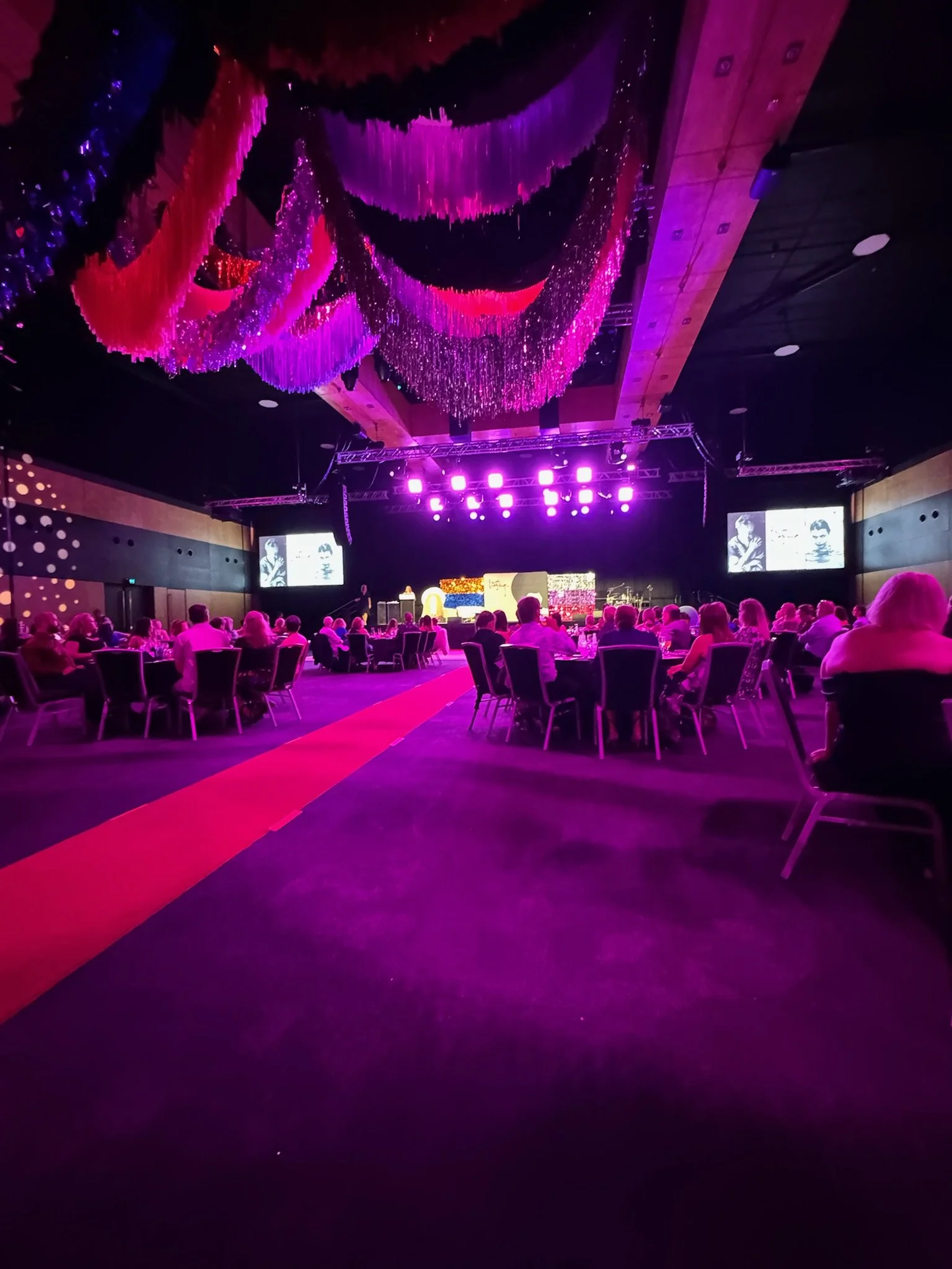 A large event hall decorated with pink, purple, and red hanging fabric and lights, with round tables set for a banquet, overlooking a stage with bright purple lighting and large screens.