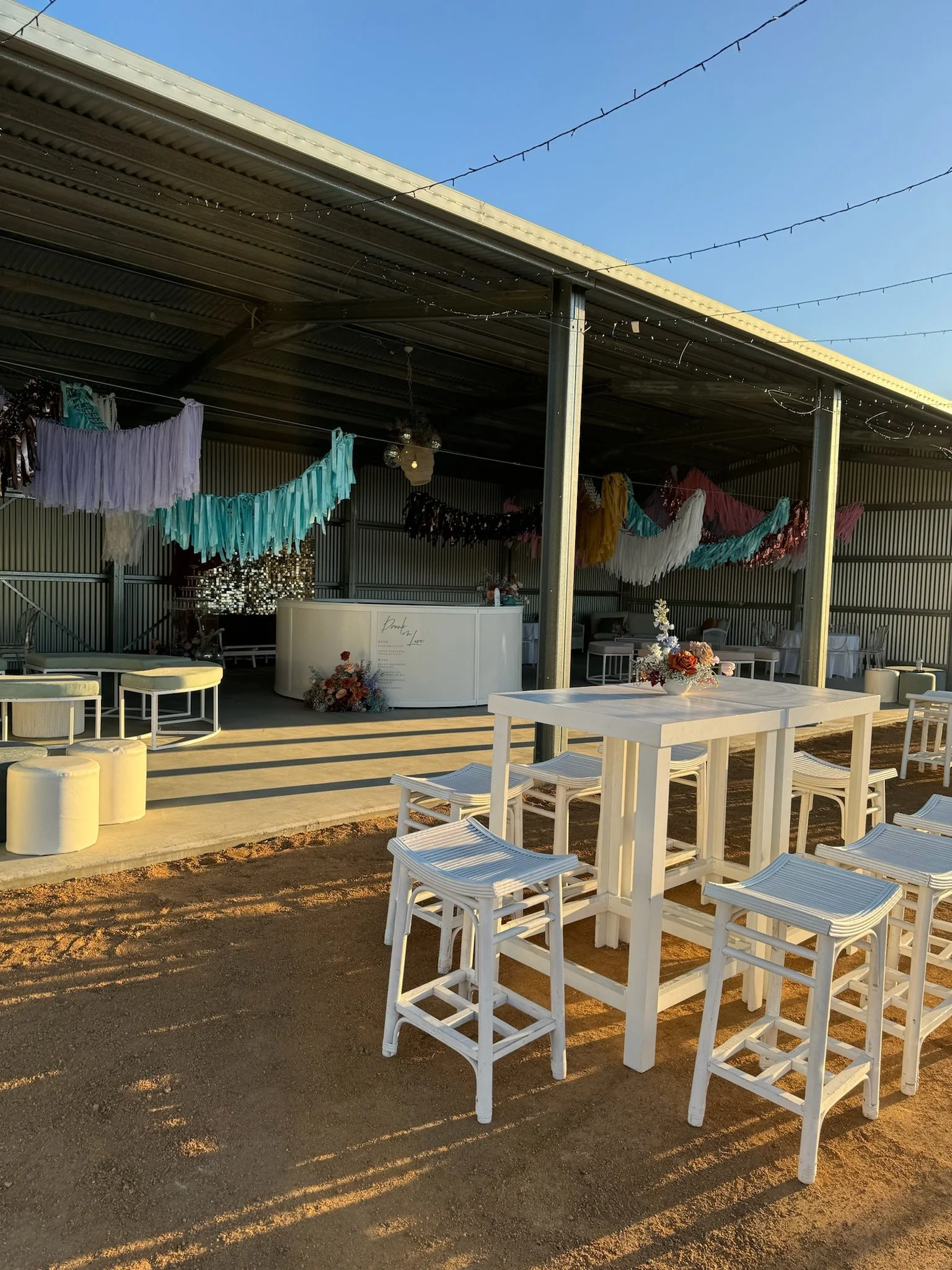 Outdoor event space decorated with white tables and stools, floral centerpieces, colorful tassel garlands hanging from a metal roof, and a bar in the background with flowers, under clear blue skies.