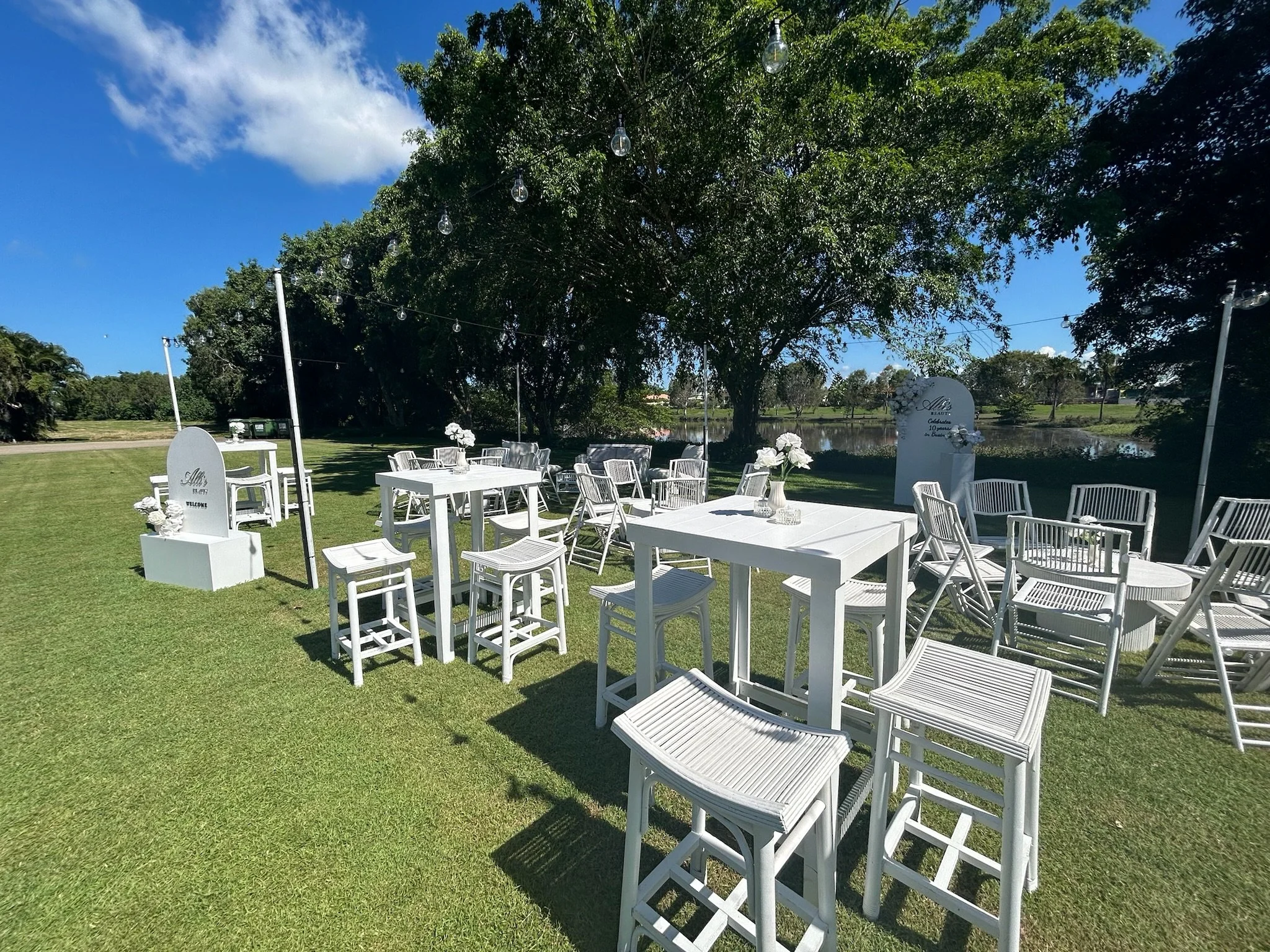 Outdoor wedding setup with white tables, chairs, and floral decorations under a large tree on a grassy area near a lake, with string lights hanging overhead.