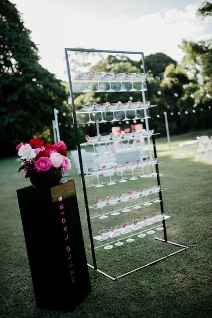 A tiered clear acrylic display stand with pink cupcakes on each shelf, outdoors on a grassy area with trees in the background. A black pedestal with a bouquet of pink roses and a sign reading 'HELLO LOVE' is next to the display.