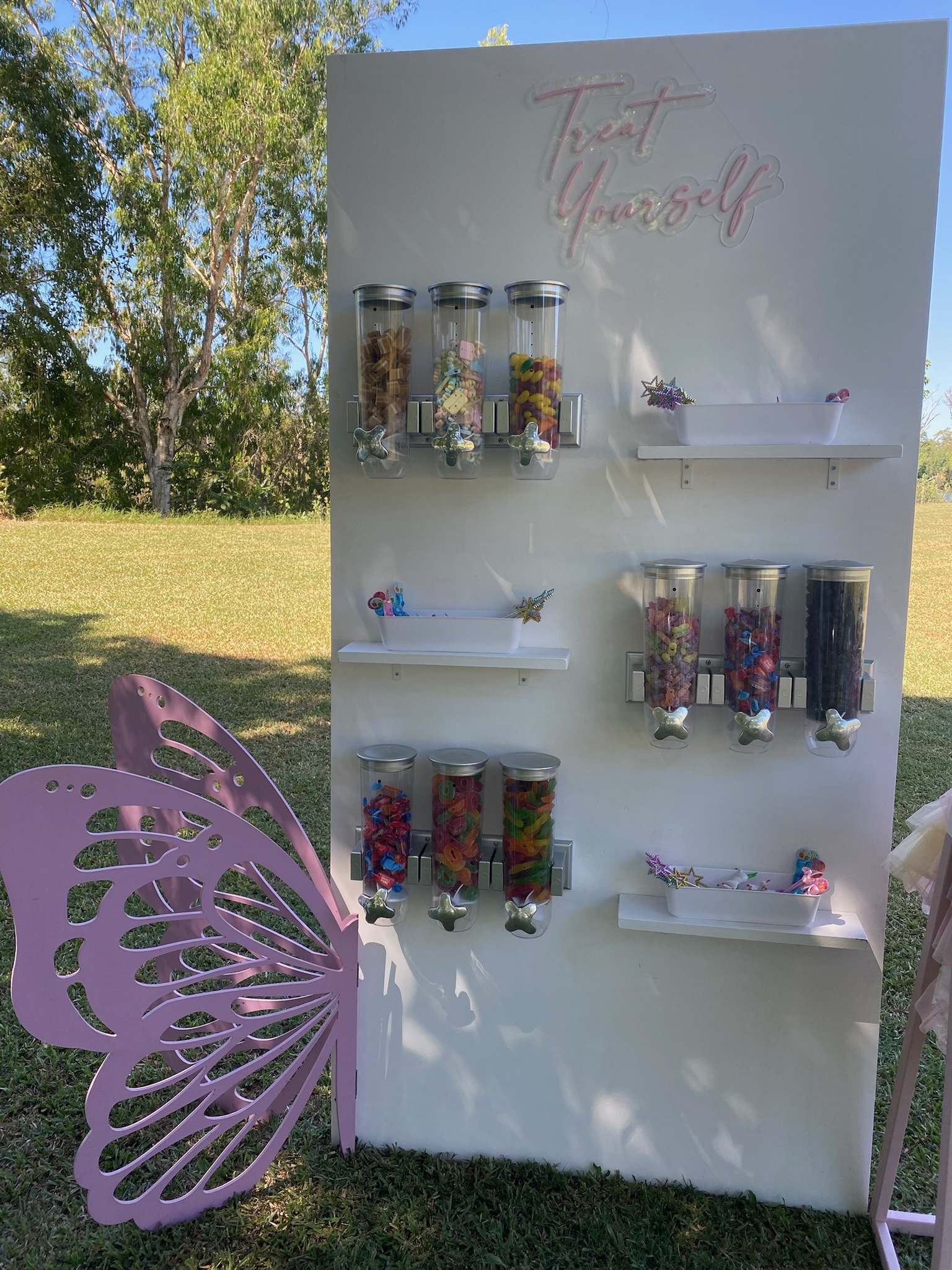 Overflowing candy station with jars of colorful candies and a neon sign that says 'Treat Yourself' at an outdoor event.