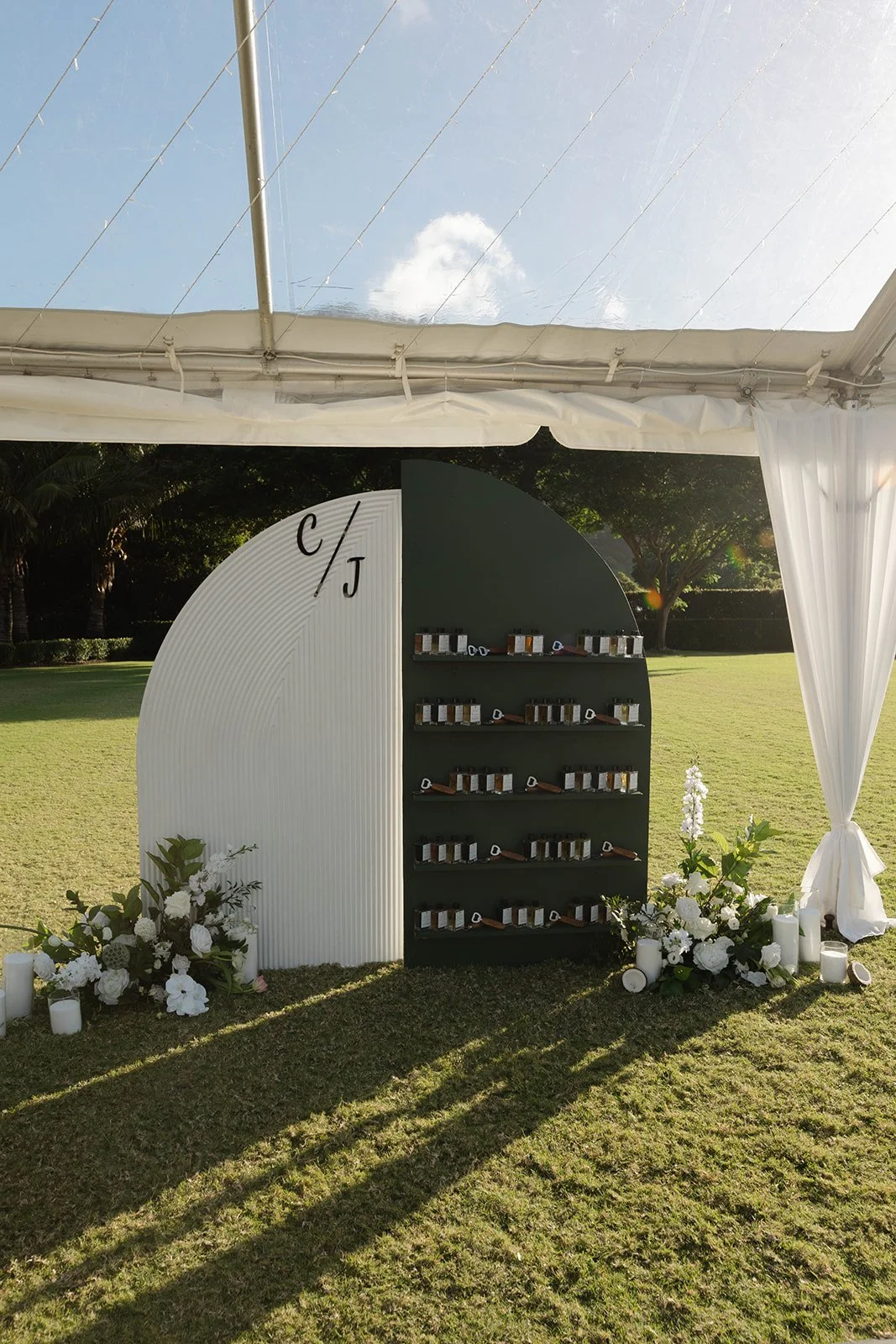 Outdoor floral display with white and green flowers and candles on the grass in front of a black and white display with shelves of small bottles, under a white canopy with clear plastic top, on a sunny day.