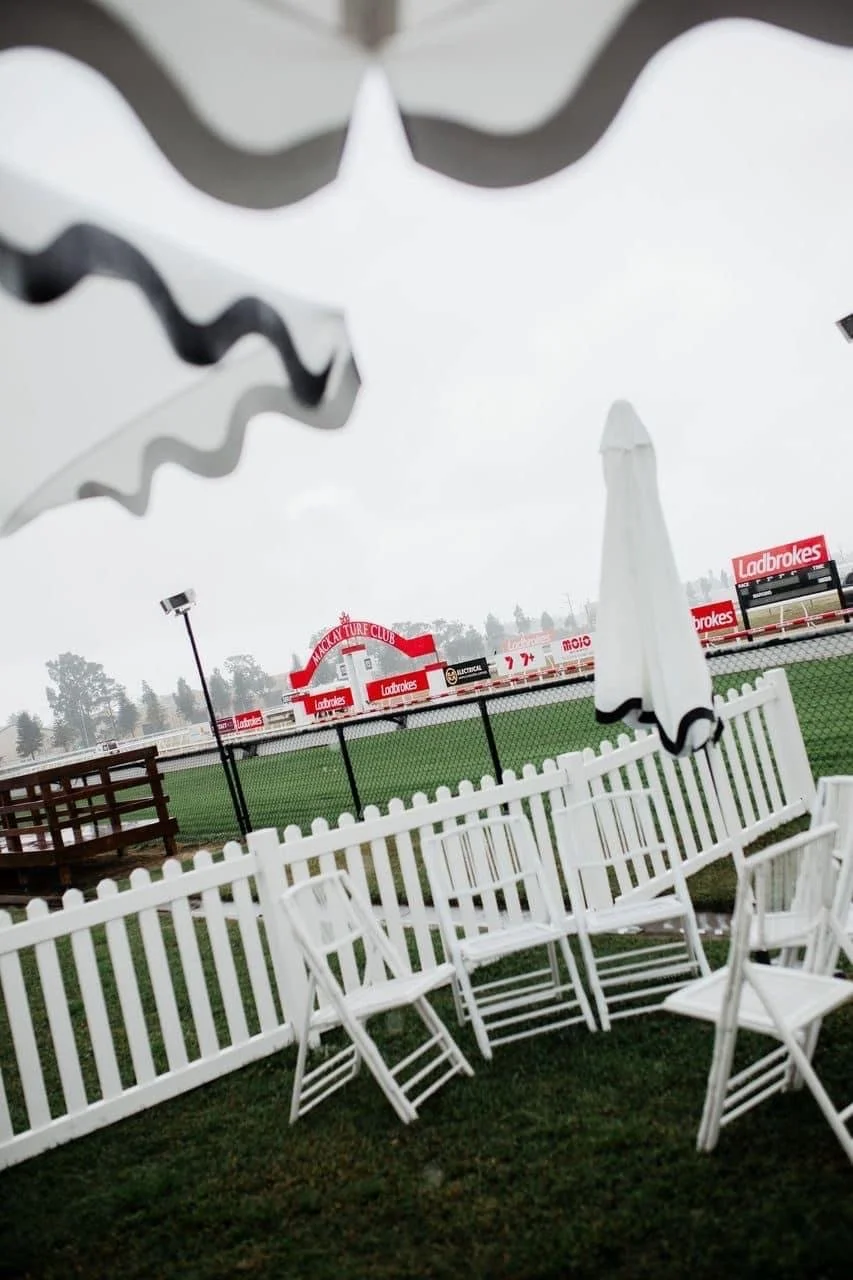 Outdoor seating area with white chairs and umbrellas facing a racetrack with advertising signs, on a rainy day.