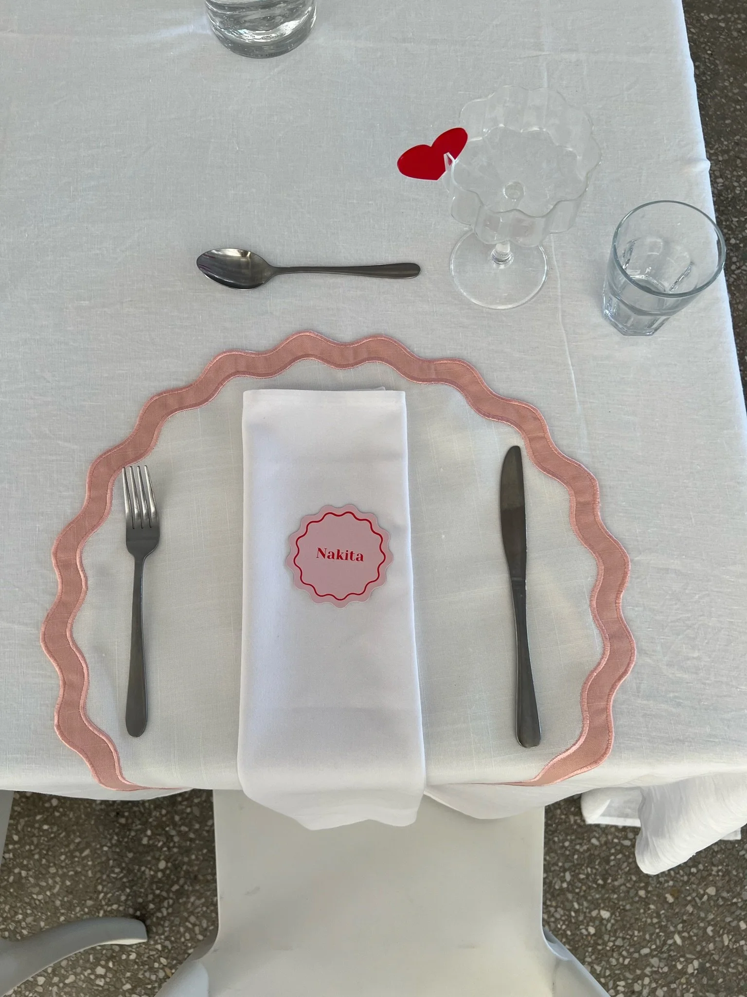 A dining table set with a white tablecloth, pink decorative placemat, white folded napkin with a name tag reading "Nakita", silverware including a fork, knife, and a spoon, a glass of water, an empty glass with a red heart-shaped sticker, and a glass