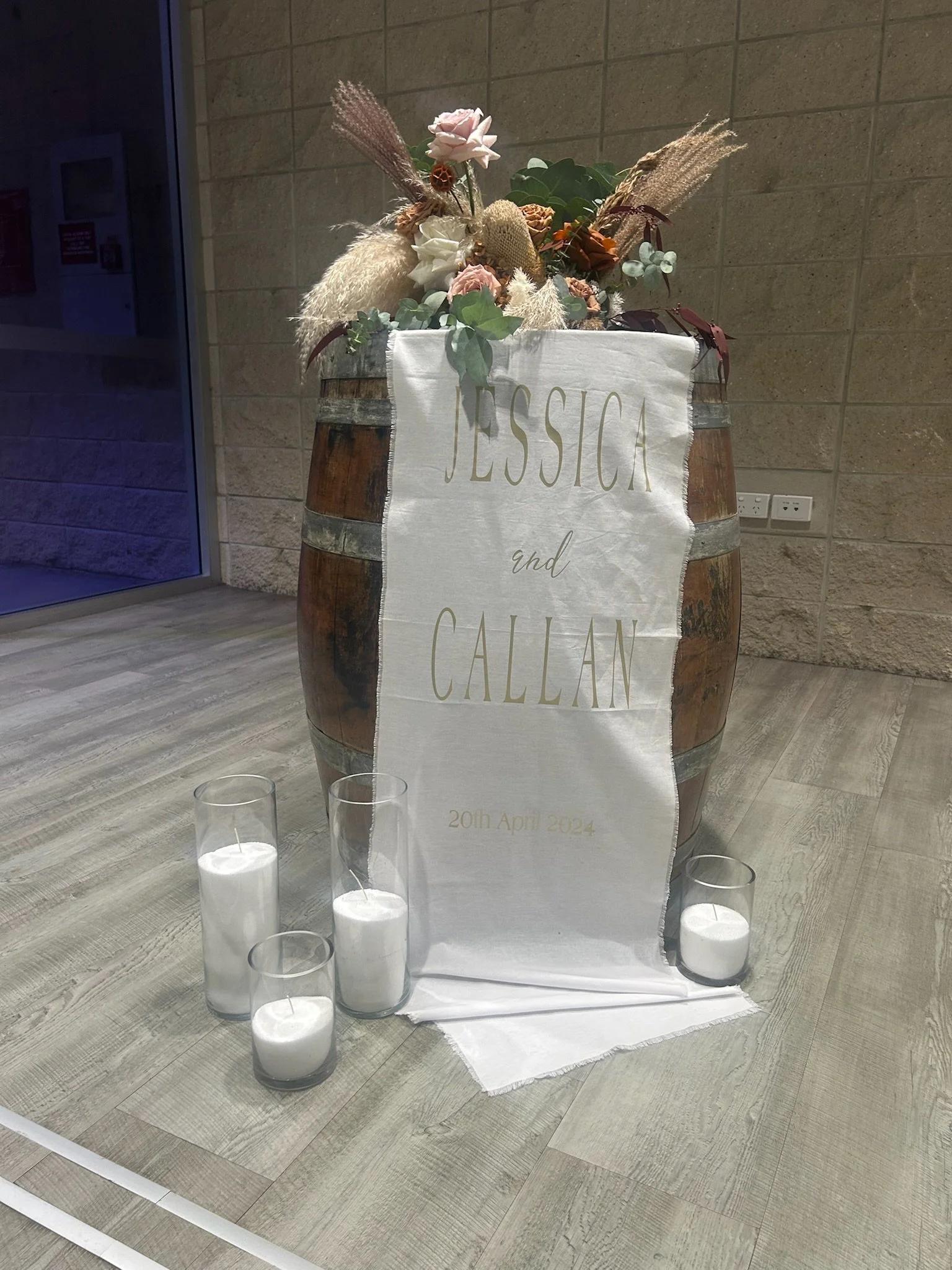 A decorative wedding display featuring a large wine barrel with flowers on top and a white fabric banner reading 'Jessica and Callan, 20th April 2024.' Four white candles in glass holders are placed in front of the barrel on a wooden floor.